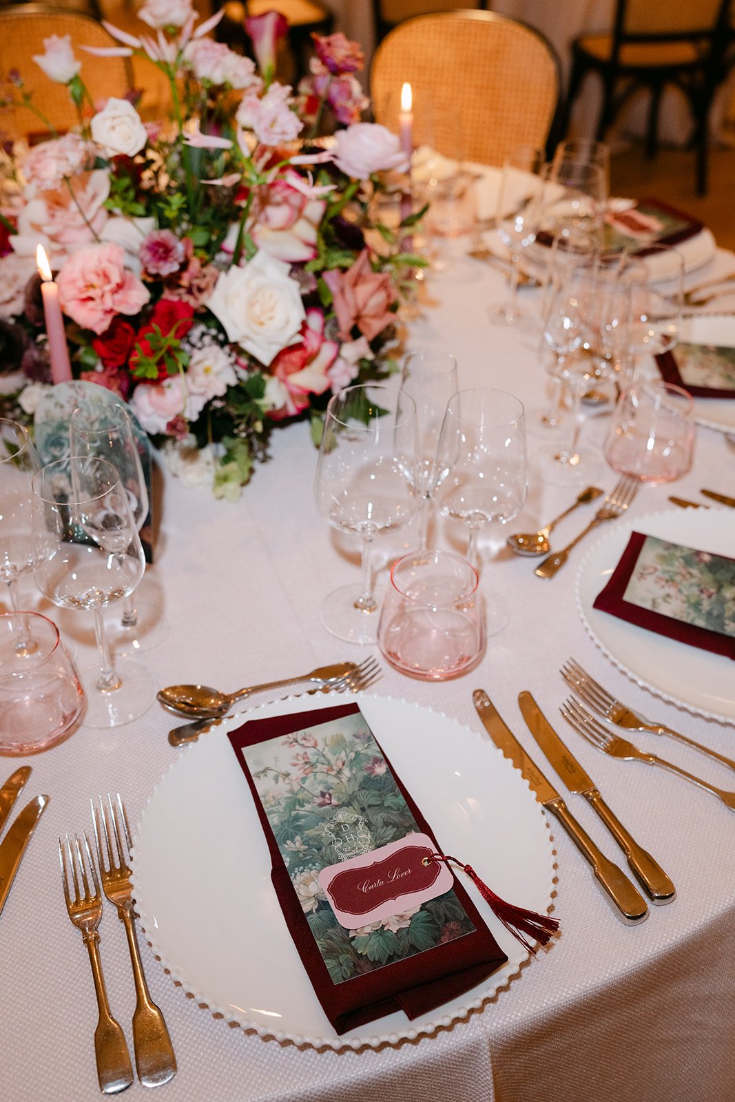 A elegantly set dining table with a large floral centerpiece, multiple wine glasses, gold cutlery. The table is decorated with a white tablecloth and candle lighting. The Botany House Wedding Florist, Kin House The Cotswolds. Planned by Get Wed
