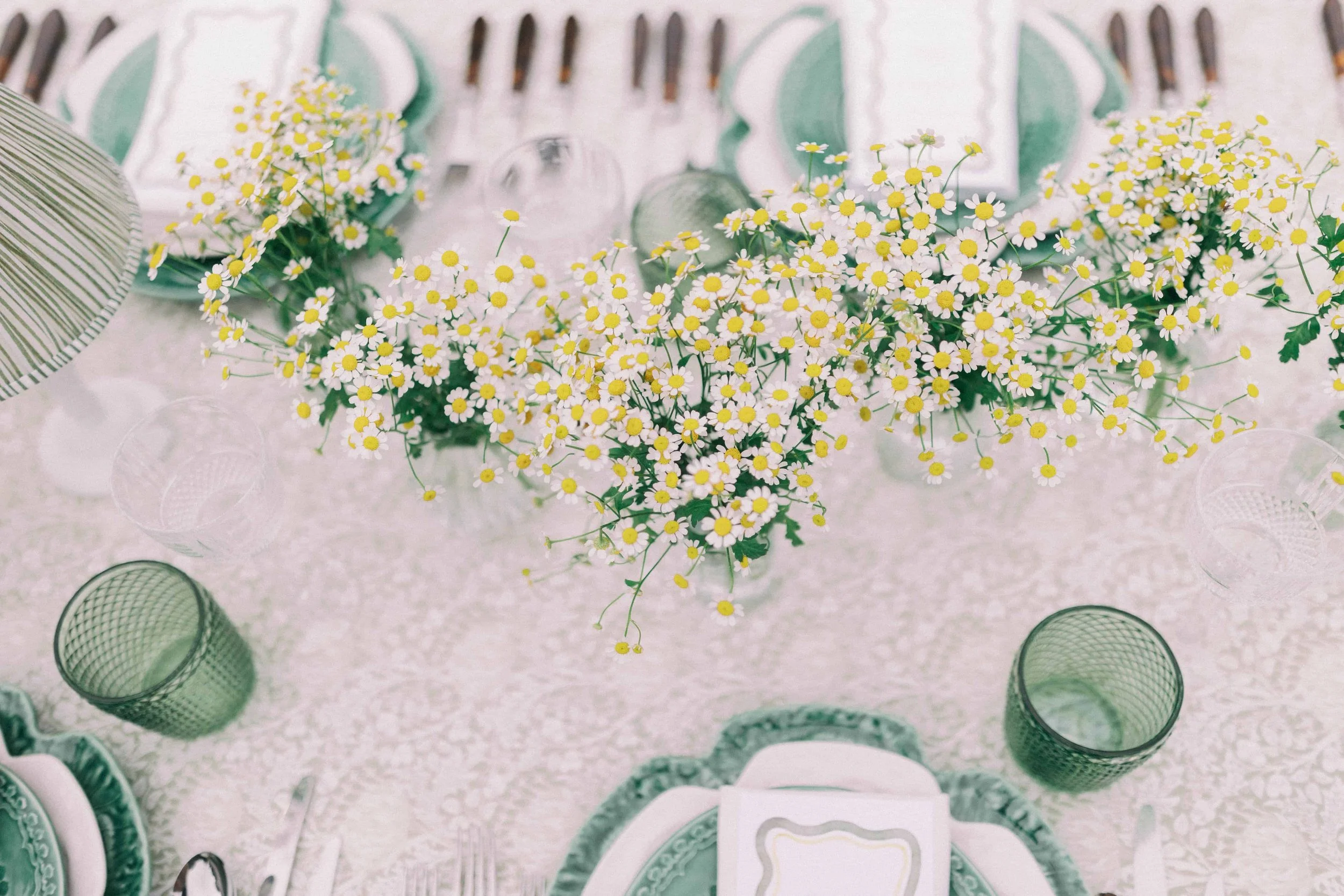 A table setting with a centerpiece of yellow and white daisies, surrounded by green and clear glass cups, plates, and silverware, on a textured cream tablecloth.