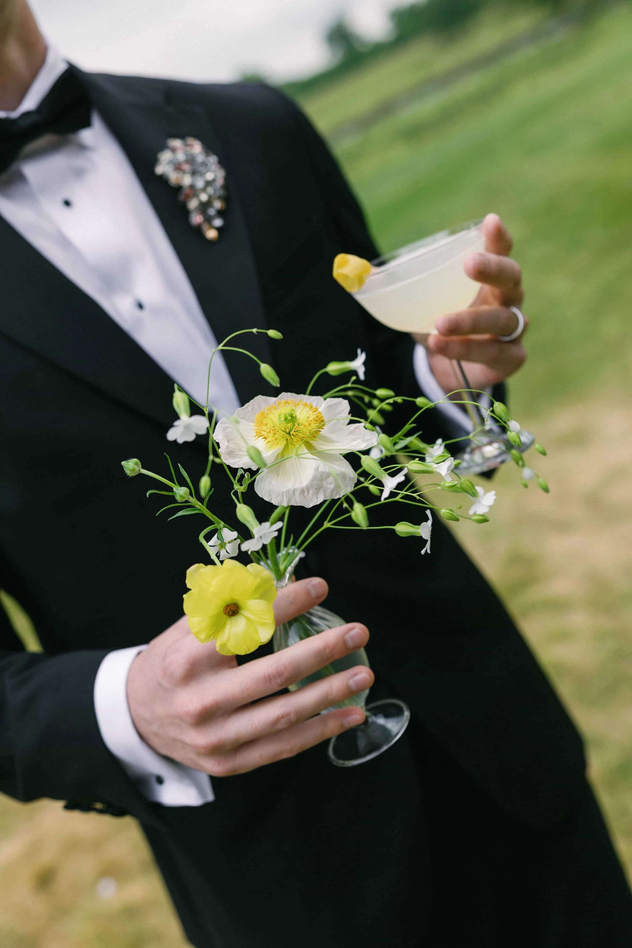 A person in a black tuxedo holding a bouquet of white and yellow flowers and a cocktail glass with a lemon wedge, outdoors with green grass in the background. The Botany House Florist at Wilderness Reserve, Planned by Attabara