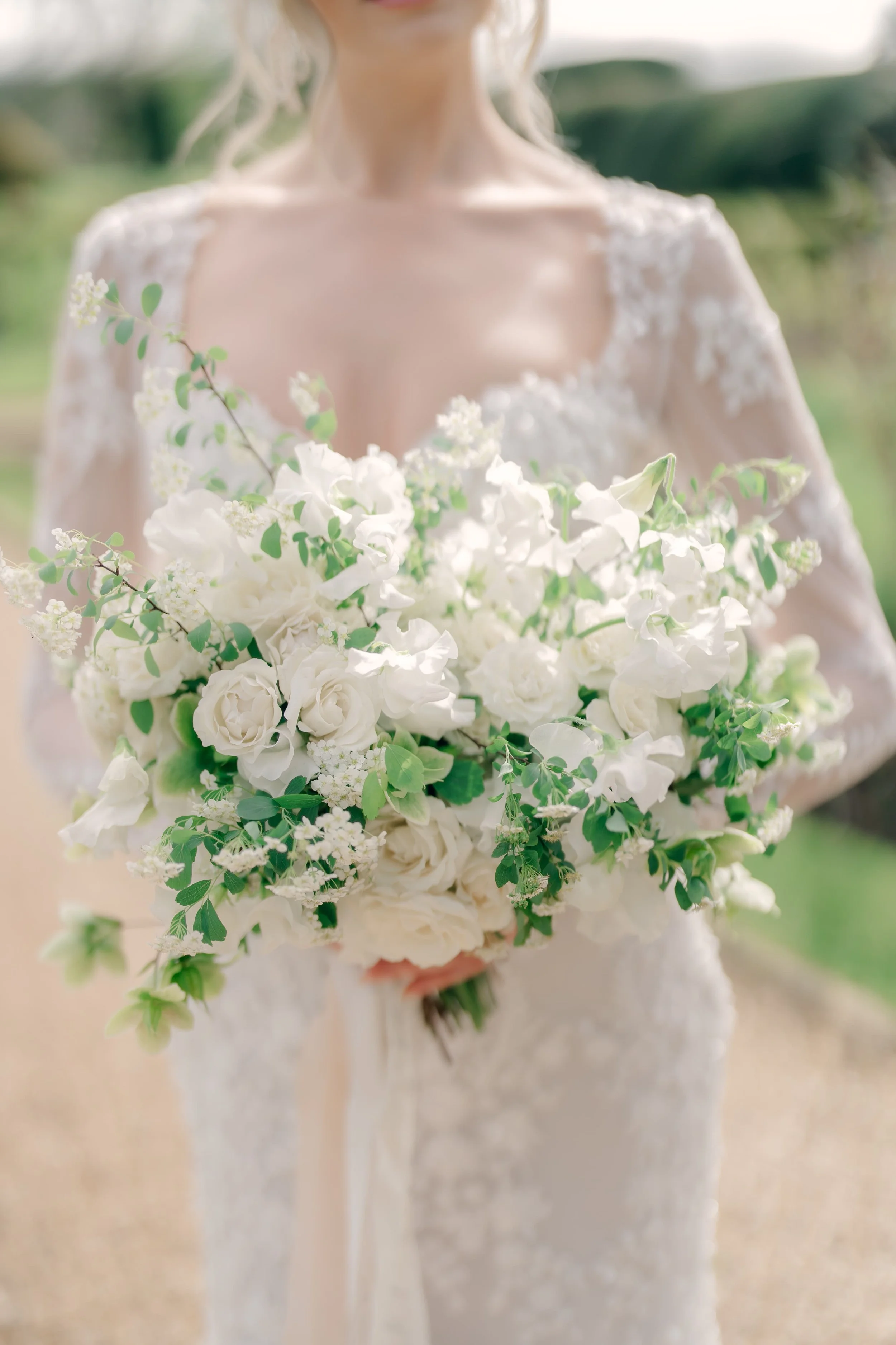 Bride holding a cascading bouquet of white roses, peonies, and greenery, wearing a lace wedding dress with lace sleeves, outdoors. Sudeley Castle The Cotswolds, Florals by The Botany House