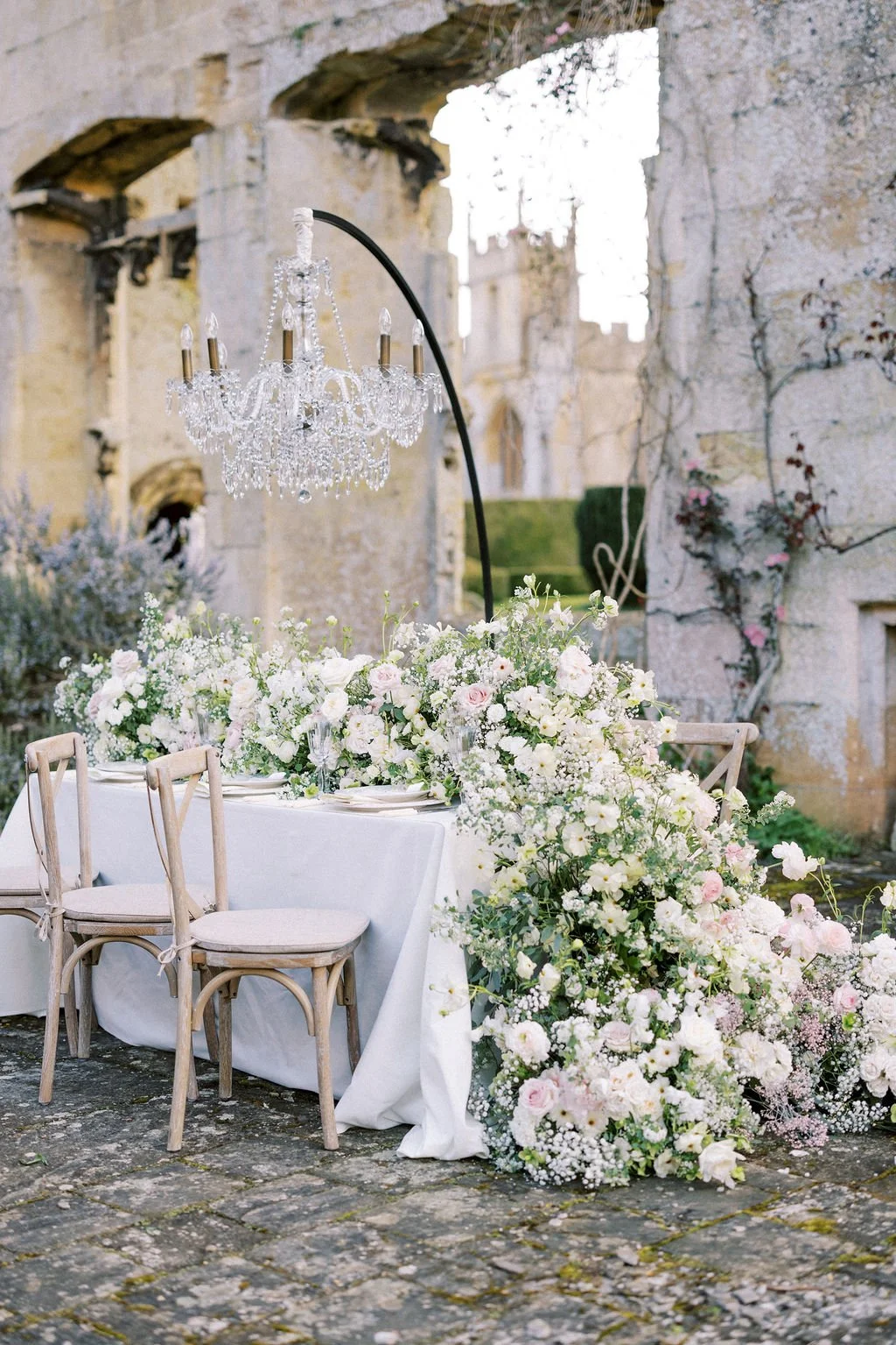Elegant outdoor dining table decorated with a lush floral arrangement, white tablecloth, and crystal chandelier hanging overhead in a rustic setting with stone walls and archways.