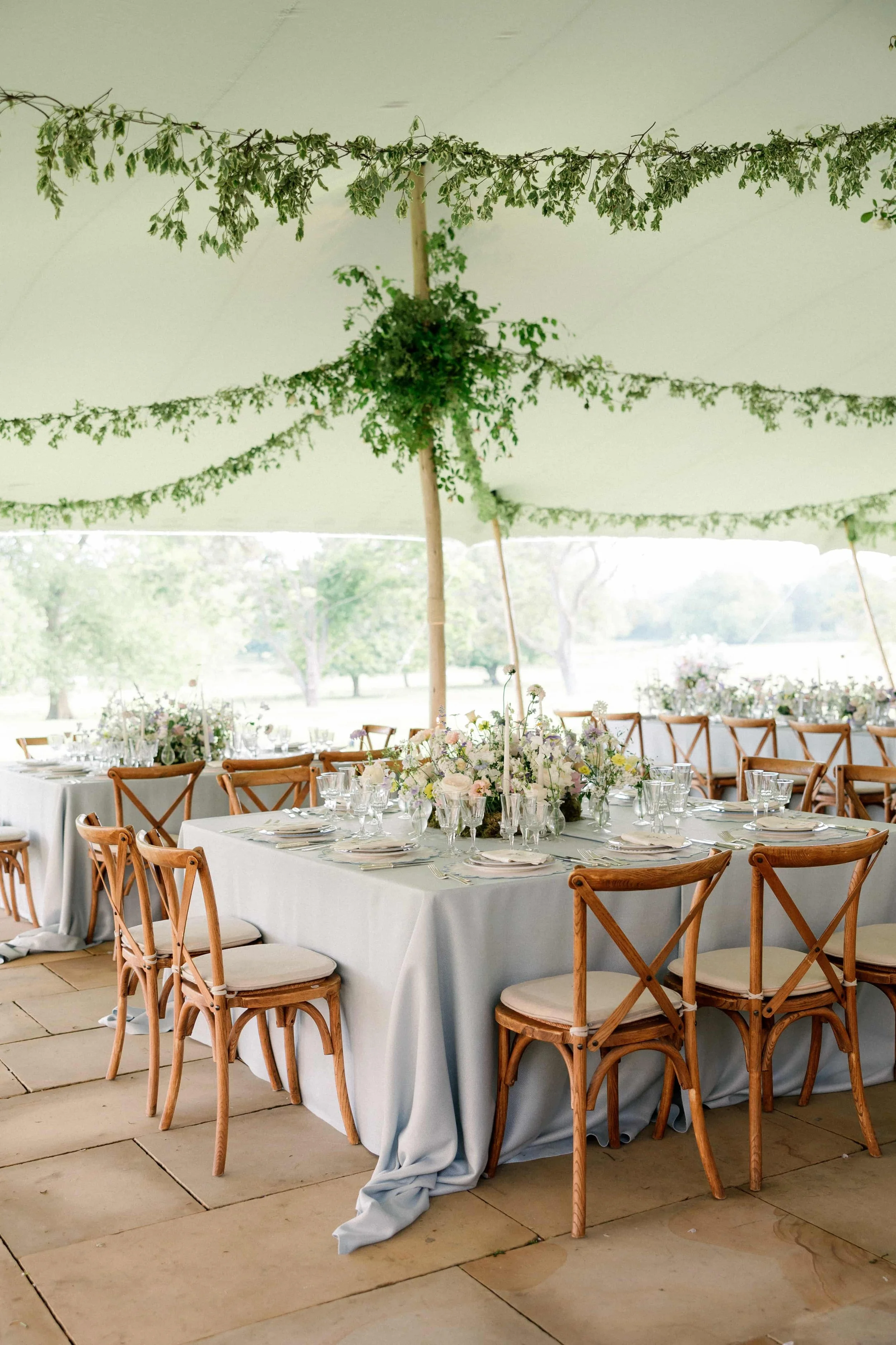 Elegant outdoor event setup under a large tent with white drapery, greenery, and floral centerpieces on a long banquet table surrounded by wooden chairs. Wilderness Reserve, Suffolk Florals By The Botany House