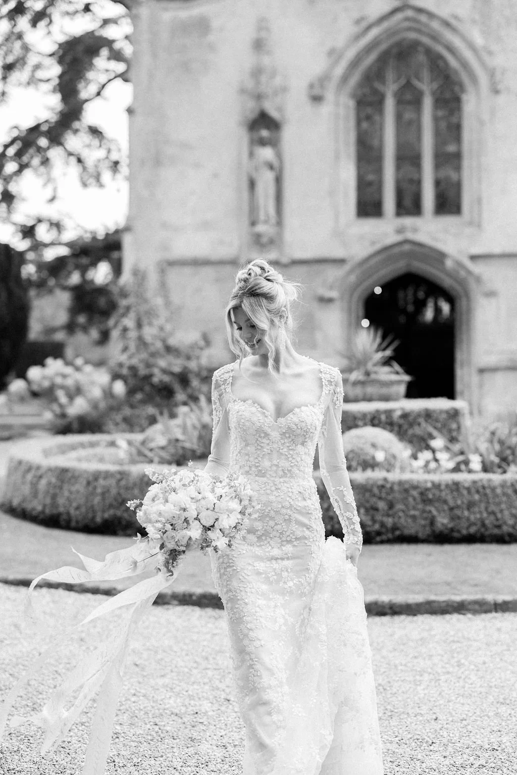 Black and white photo of a smiling bride in a lace wedding gown holding a bouquet, standing outside a church.