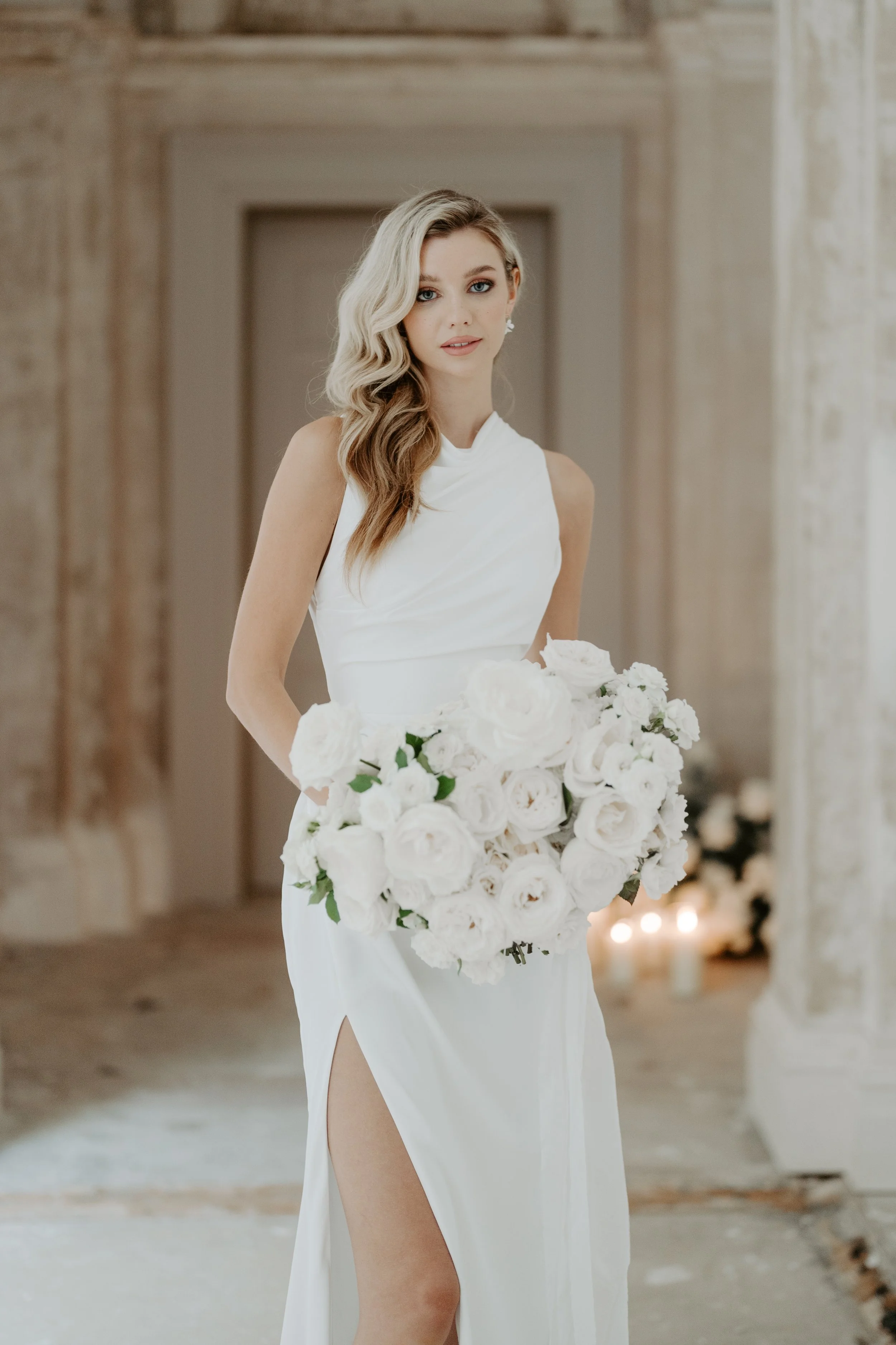 A bride in a white wedding gown holding a bouquet of white roses in an elegant, softly lit room.