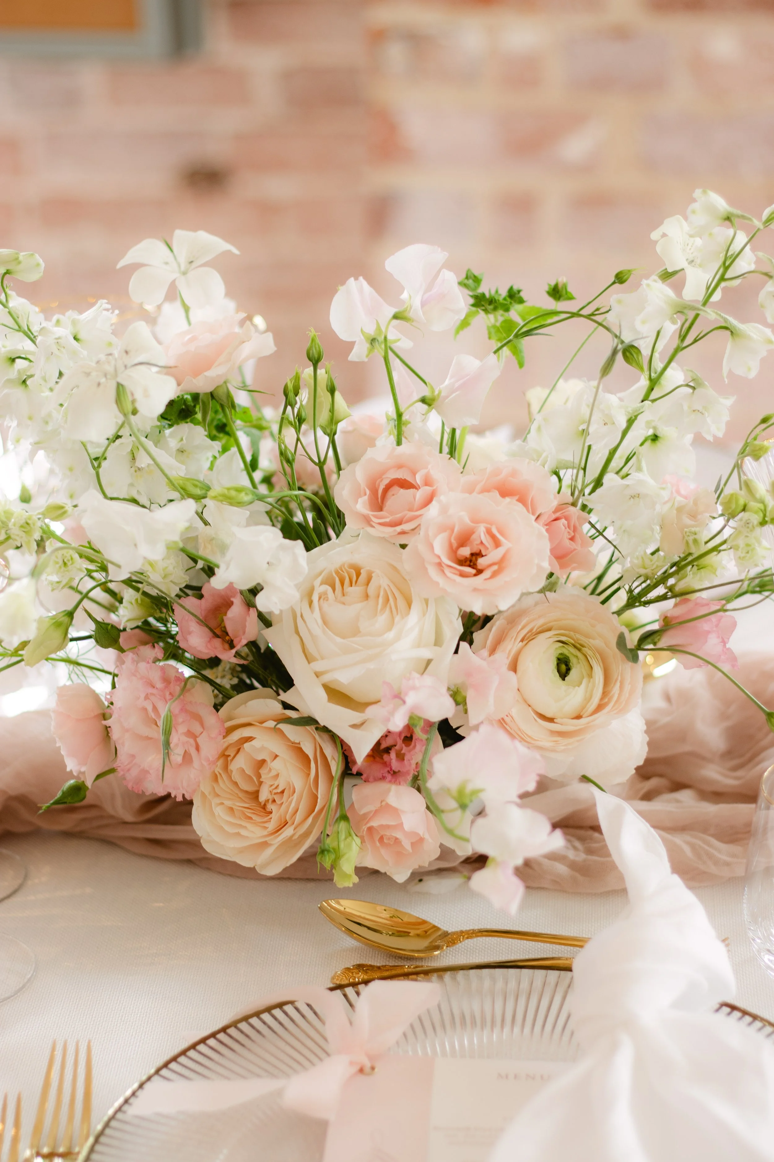 A centerpiece of pastel pink and white flowers on a table with gold cutlery and a menu card, in front of a blurred brick wall background.