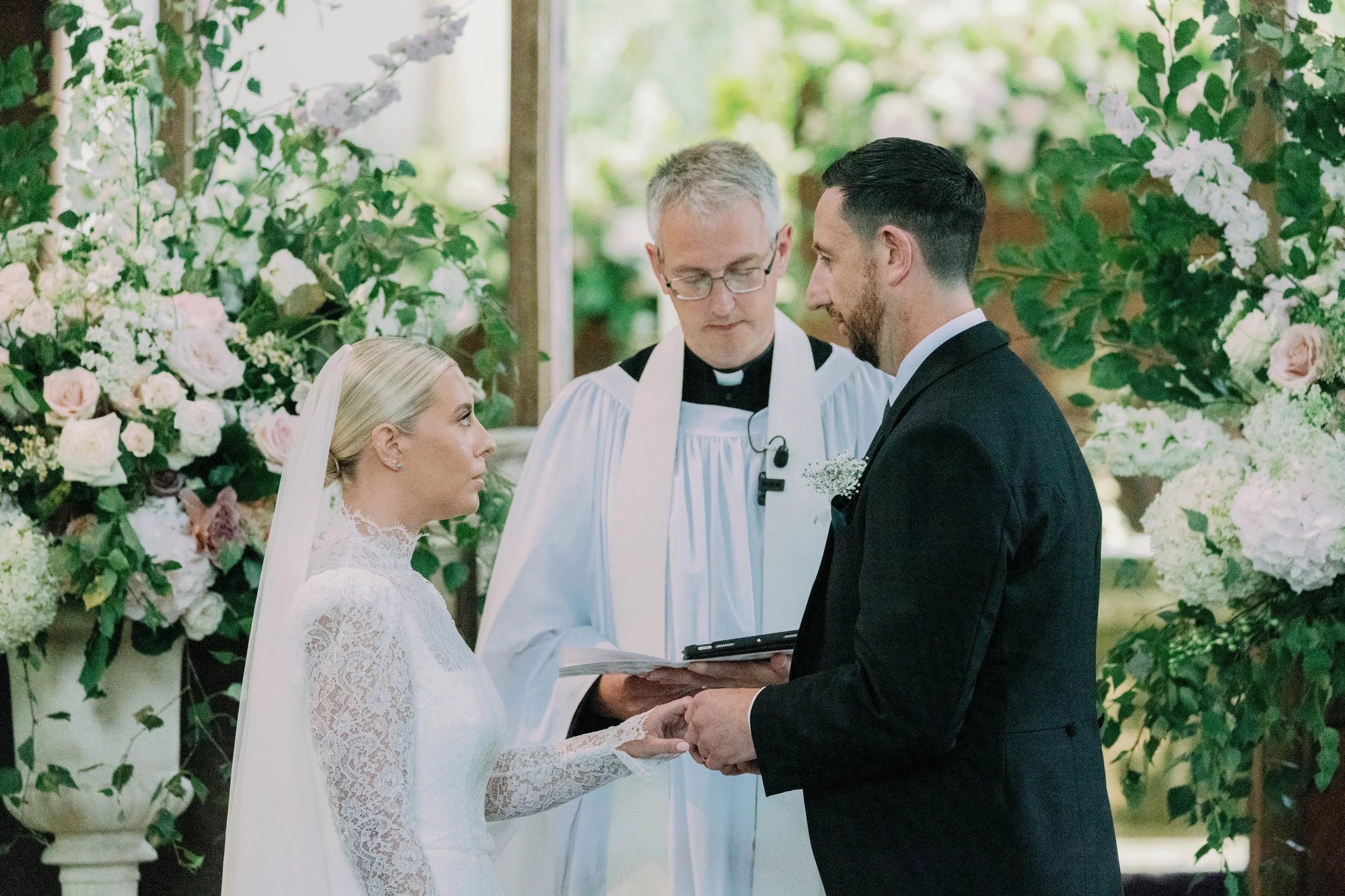 A bride and groom holding hands during their wedding ceremony in a church decorated with garden style flowers. A priest is officiating the ceremony, standing behind them with a bible.