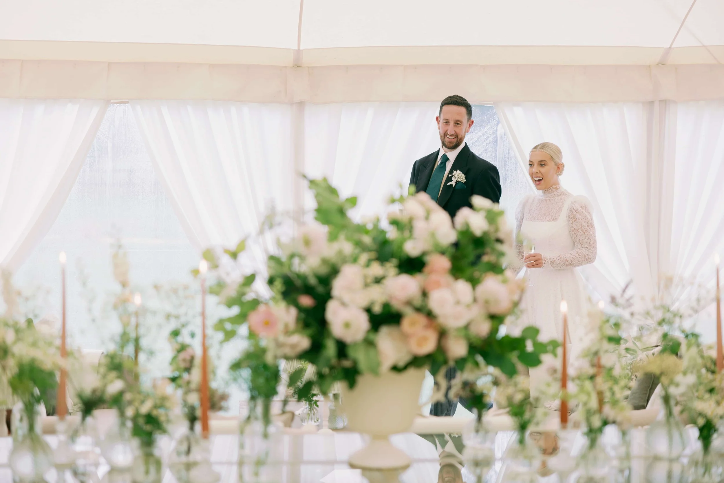 A bride and groom at their wedding reception, standing behind a table decorated with a large floral centerpiece and lit candles inside a white tent with draped curtains.