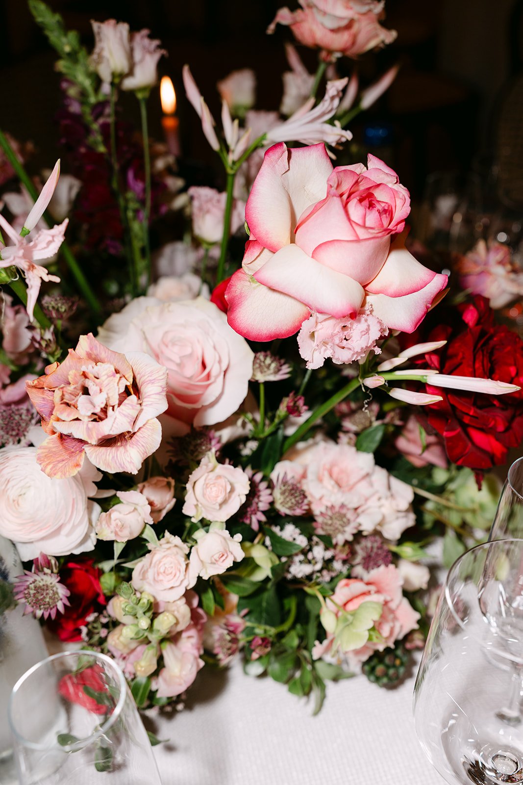 A floral arrangement featuring pink and white roses, peonies, and various other flowers on a white tablecloth, with glassware in the foreground.