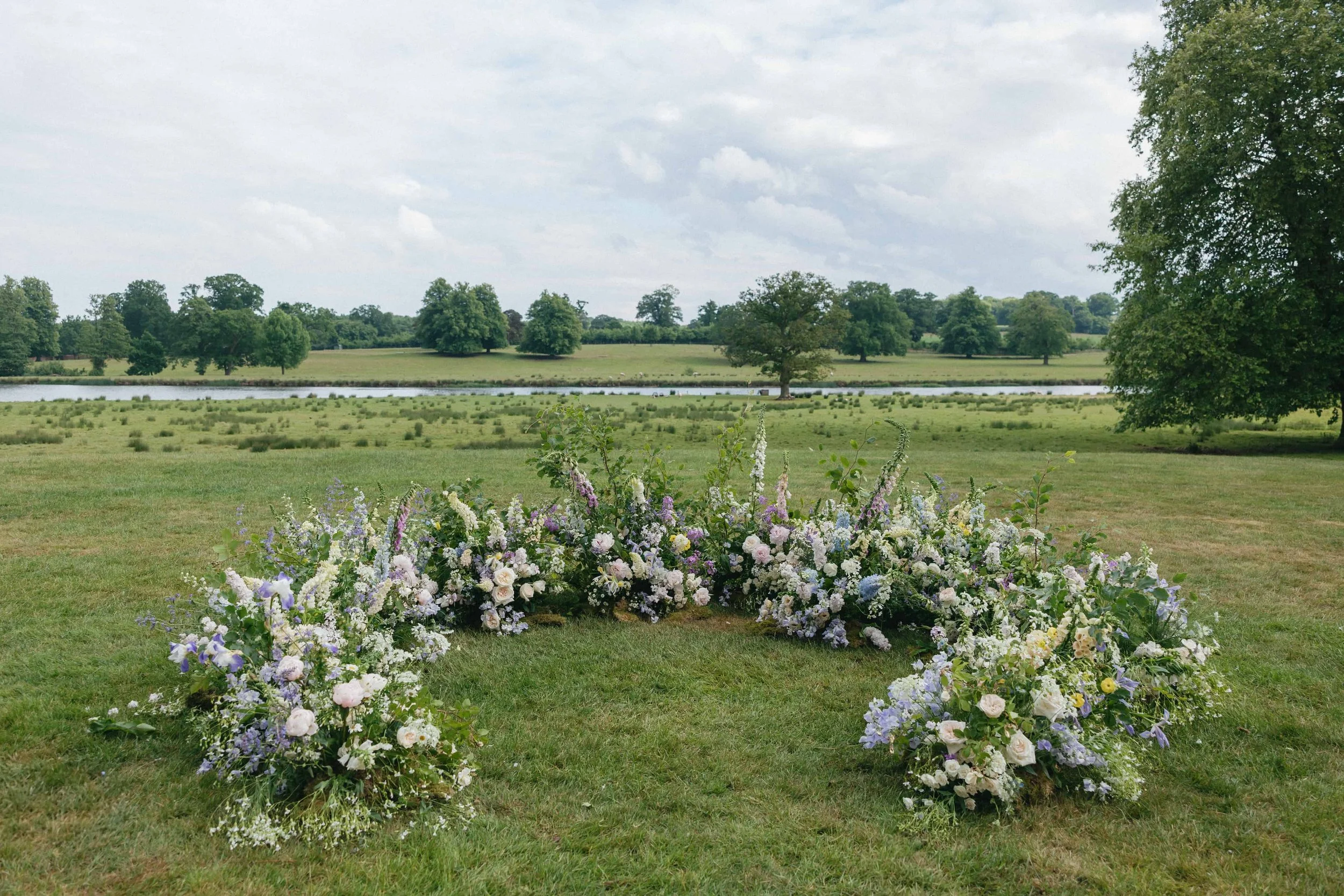 Floral arrangement in a heart shape on grass with trees and a lake in the background under a partly cloudy sky. Wilderness Reserve, Suffolk Florals By The Botany House