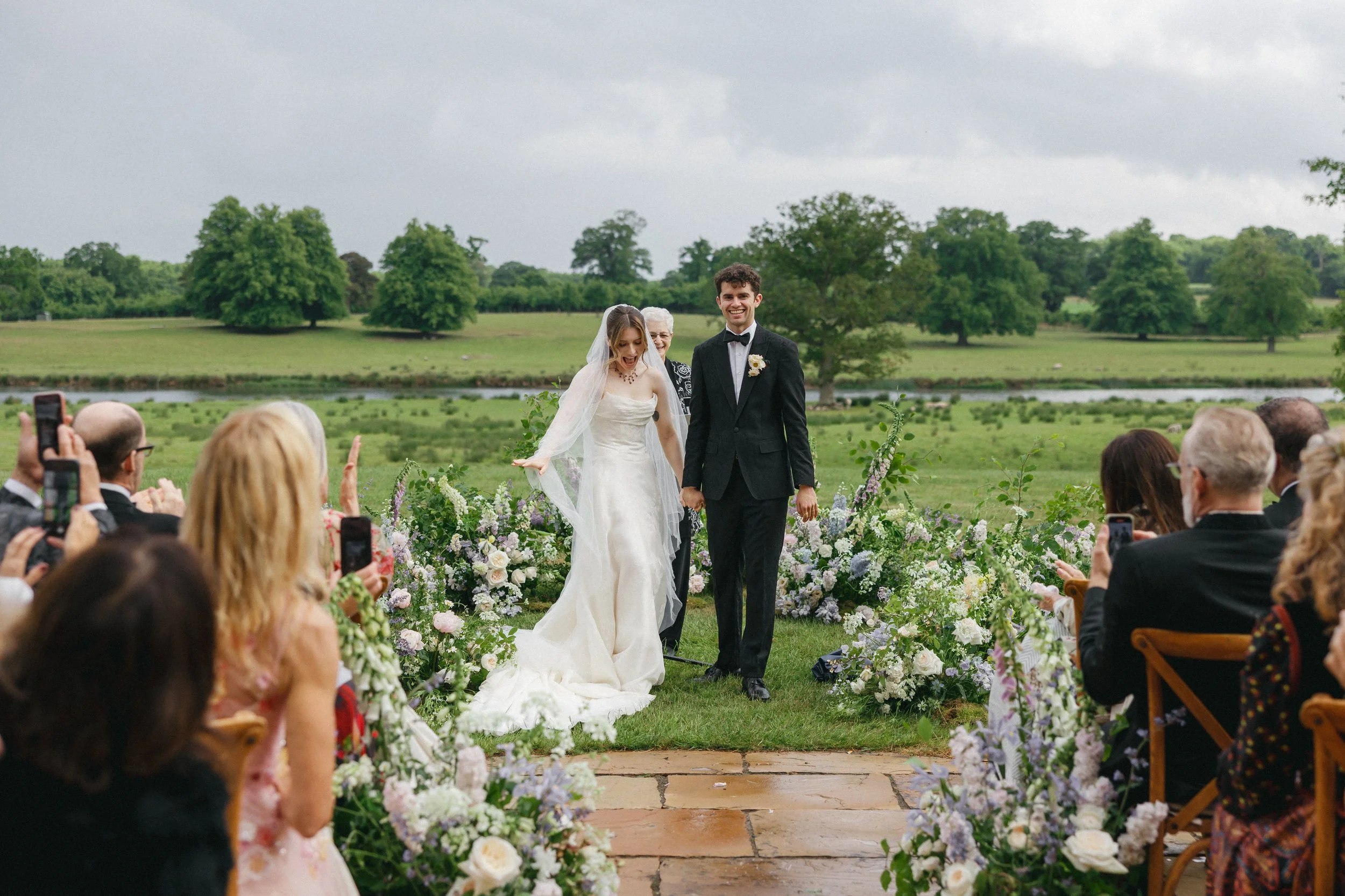 Bride and groom walking down the aisle outdoors at their wedding, surrounded by floral arrangements and guests taking photos, with a scenic green landscape and water in the background.