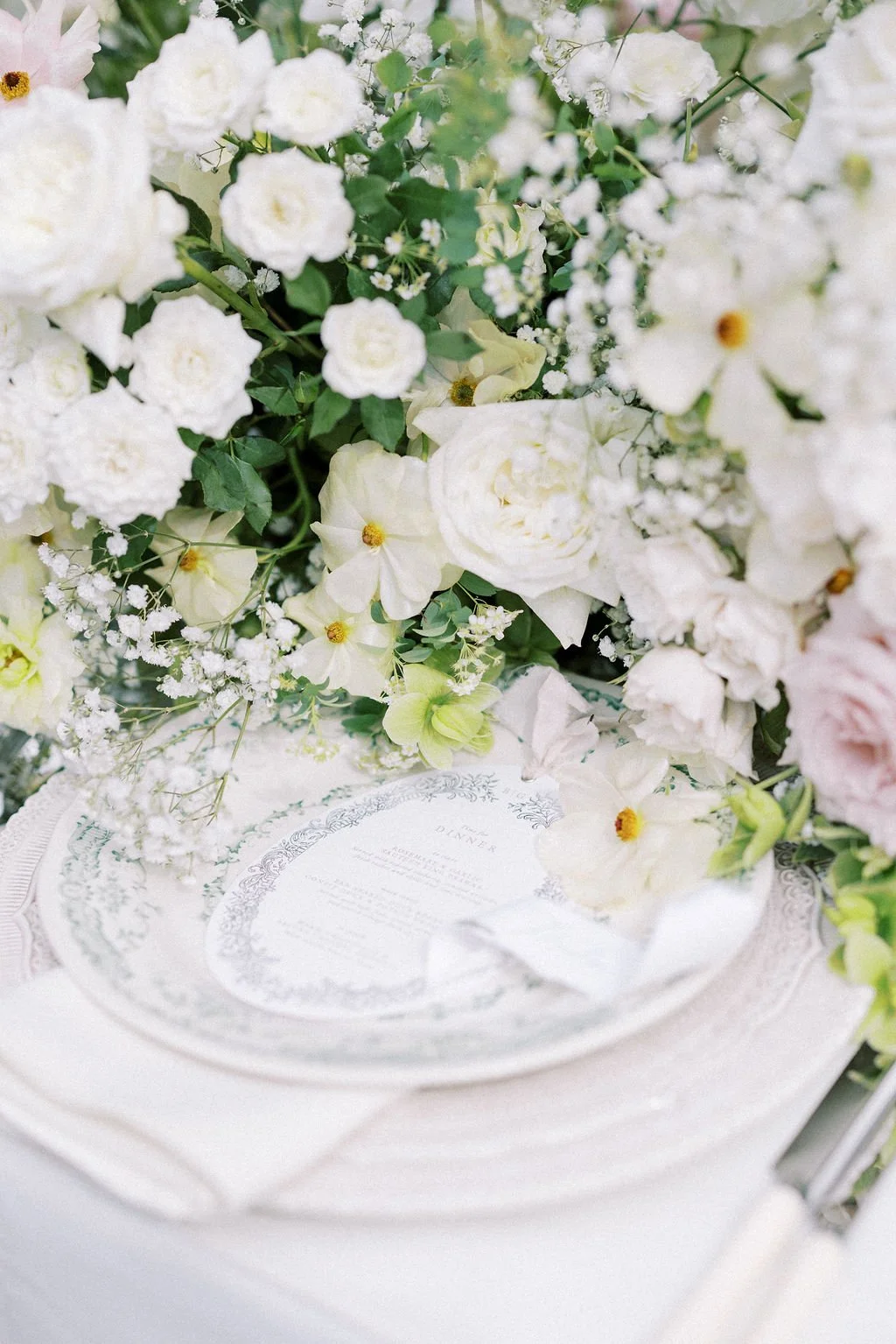 A floral centerpiece with white flowers and greenery on a table with a decorative plate and menu.