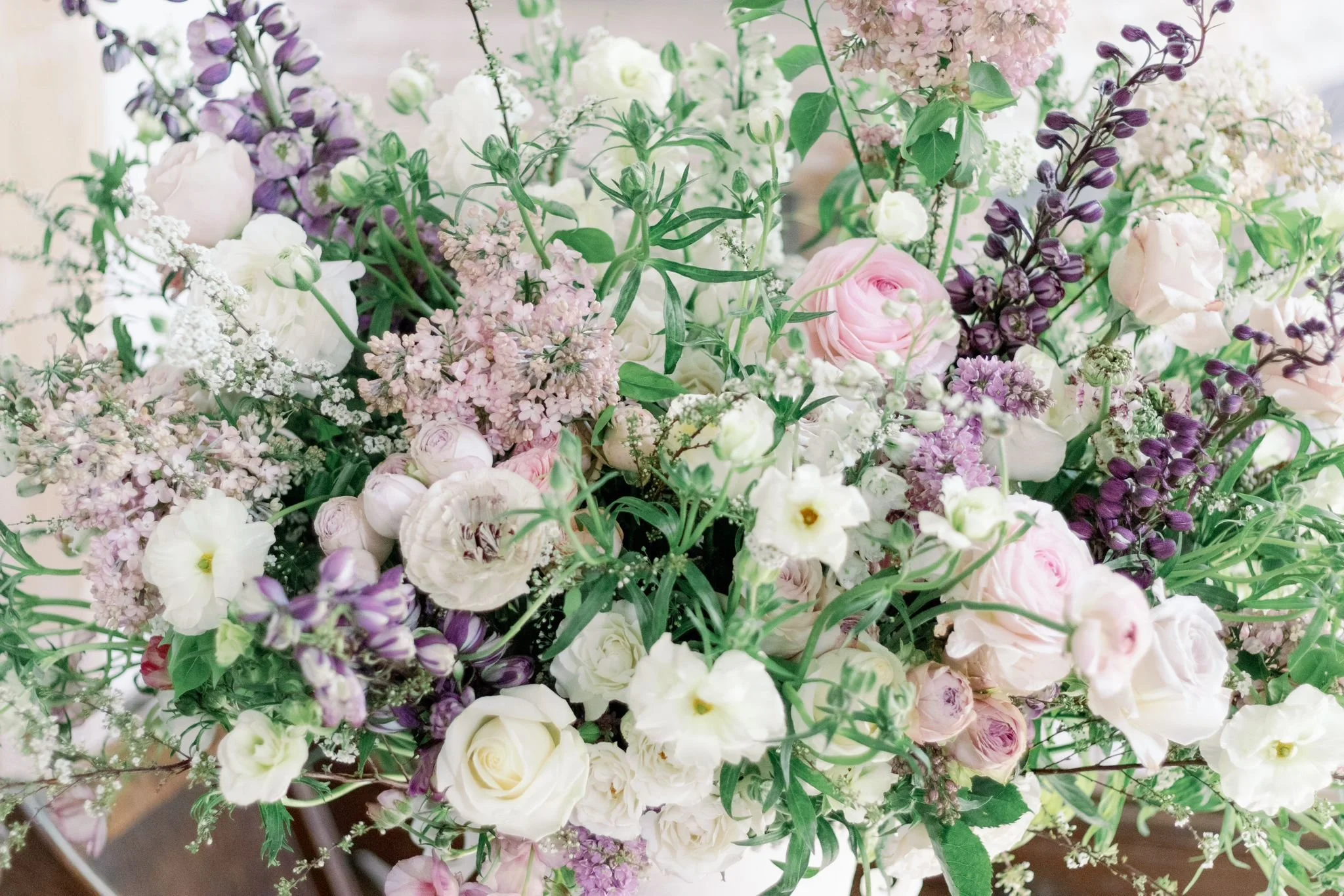 A large floral arrangement of white, pink, and purple flowers, including roses, lisianthus, stock, and other mixed florals, with green foliage.