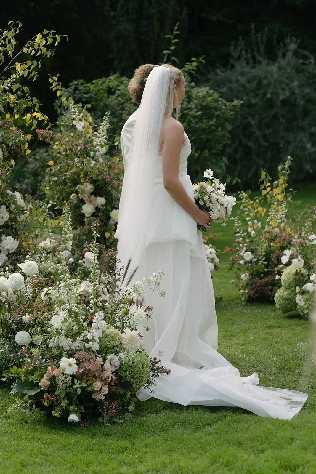 Bride holding a seasonal white bouquet at a Cornwell Manor wedding, surrounded by luxury garden-style floral installations by The Botany House, best luxury wedding florist.