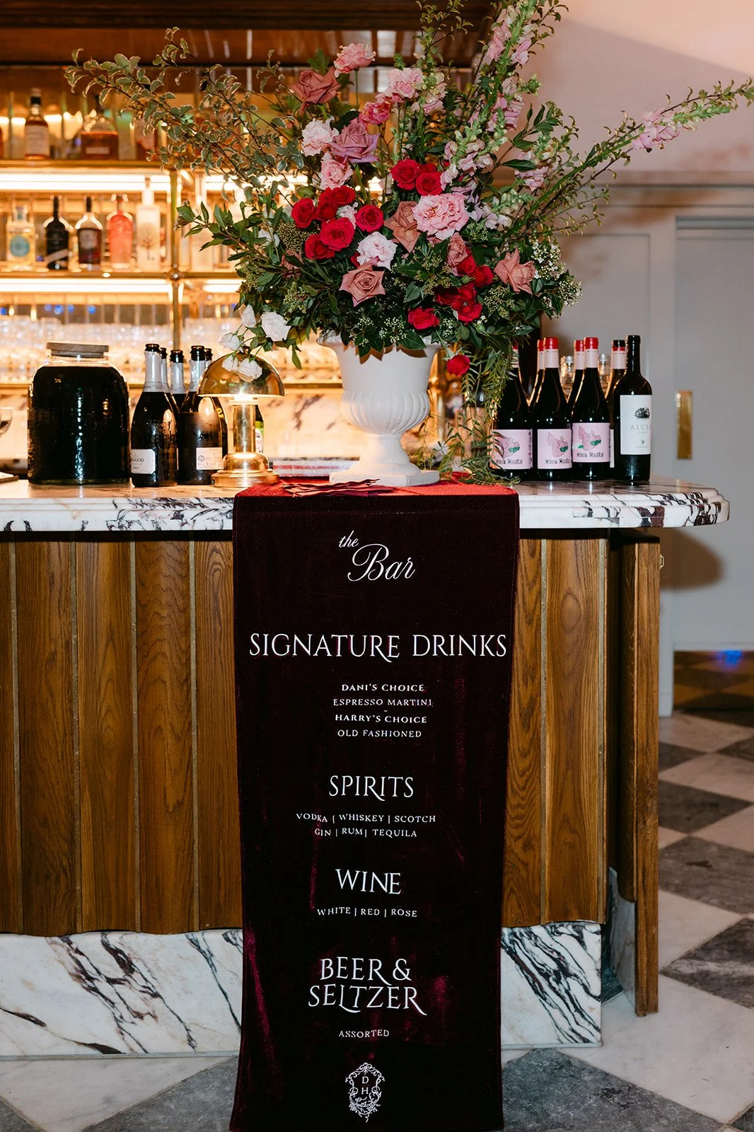 A bar counter with a large floral arrangement in a white vase, bottles of wine, and a black Erica sign displaying signature drinks, spirits, wine, and beer options.
