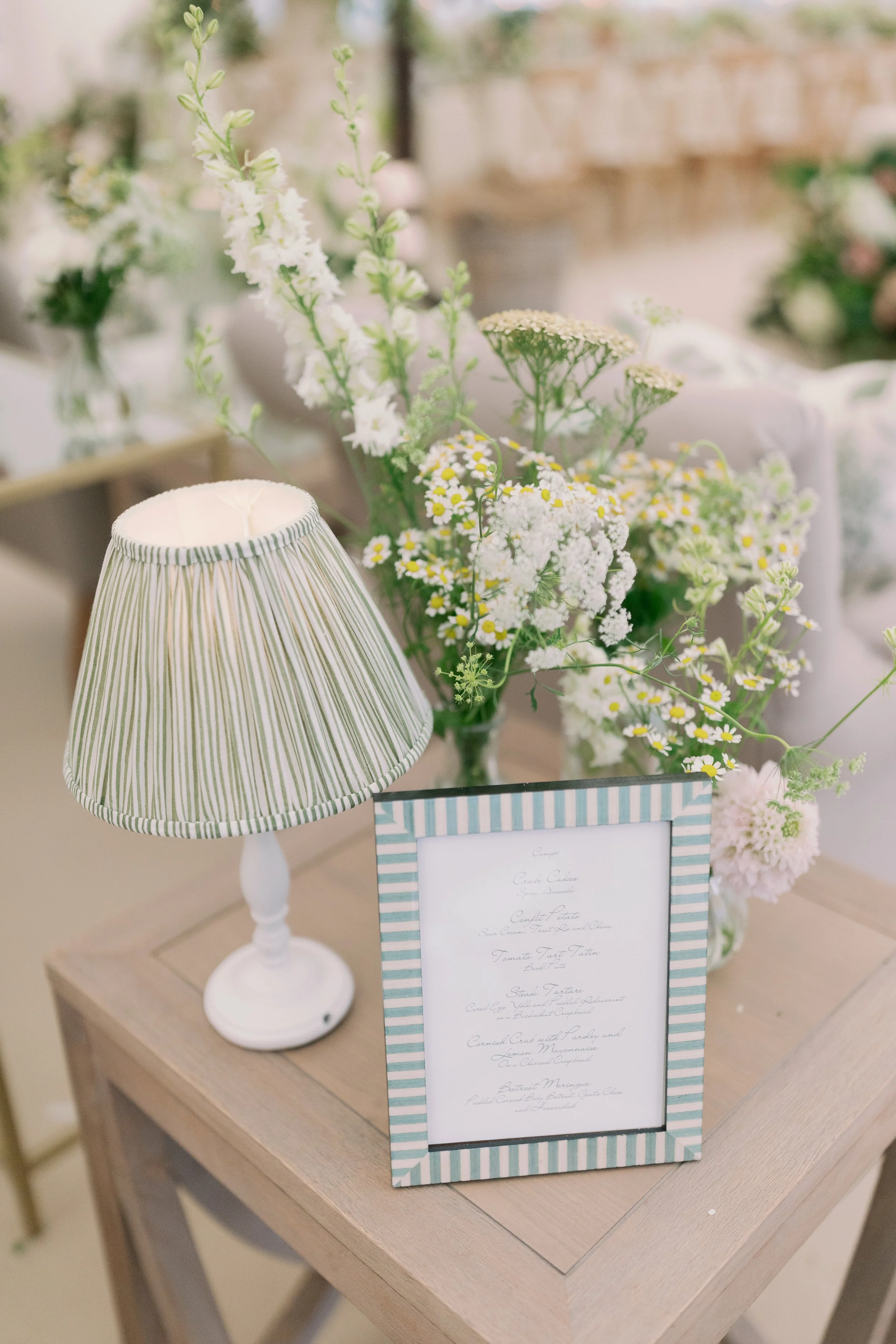 A small white table with a striped lamp, a framed menu, and a bouquet of daisies in a glass vase.