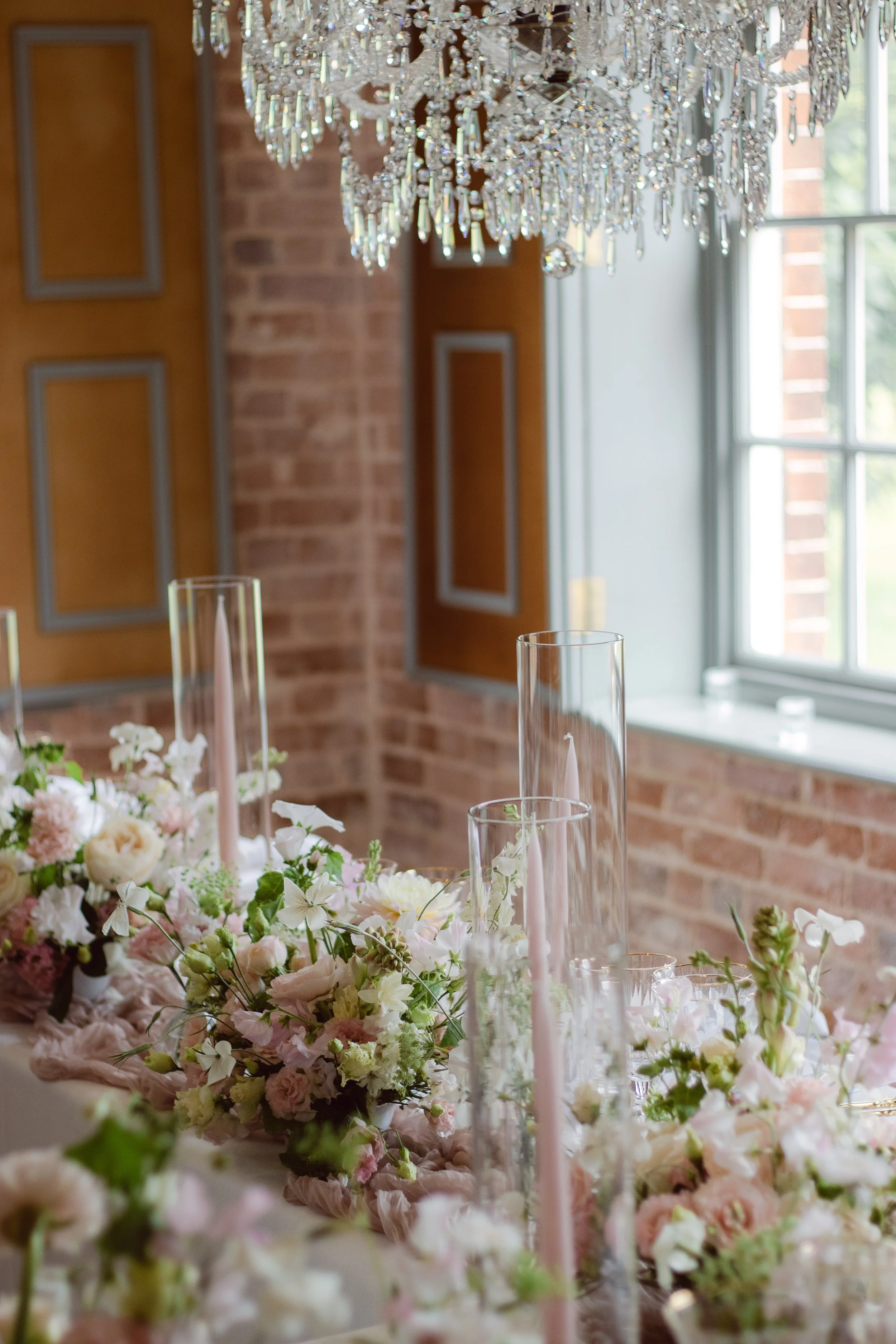 Elegant dining table with pink and white floral arrangements, tall glass candle holders, and a crystal chandelier in a decorated event space with brick walls and windows.
