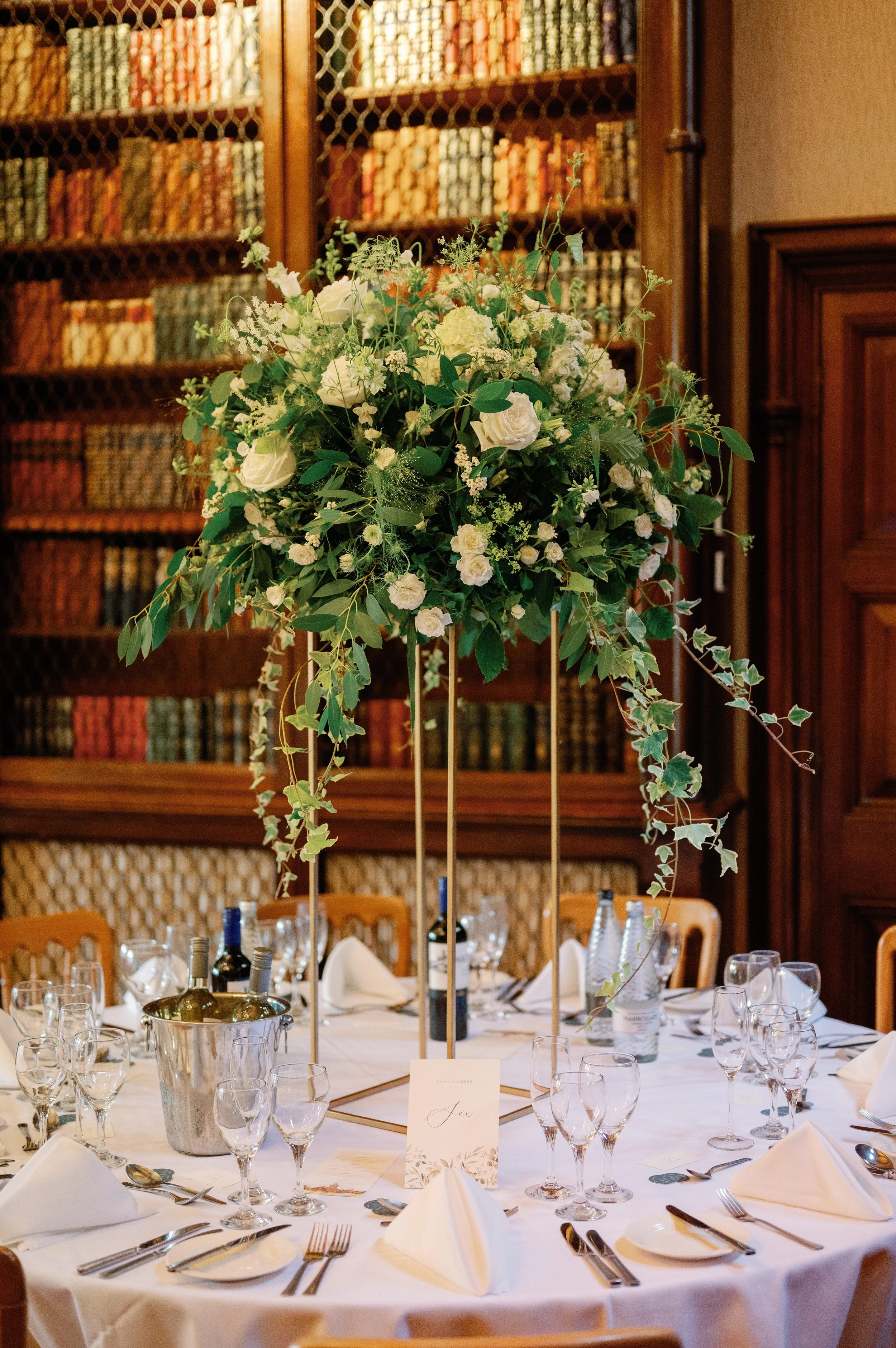Elegant round table decorated with a tall floral arrangement in a gold stand, surrounded by wine glasses, water bottles, and white napkins, set in a wood-paneled room with a bookshelf filled with books in the background.