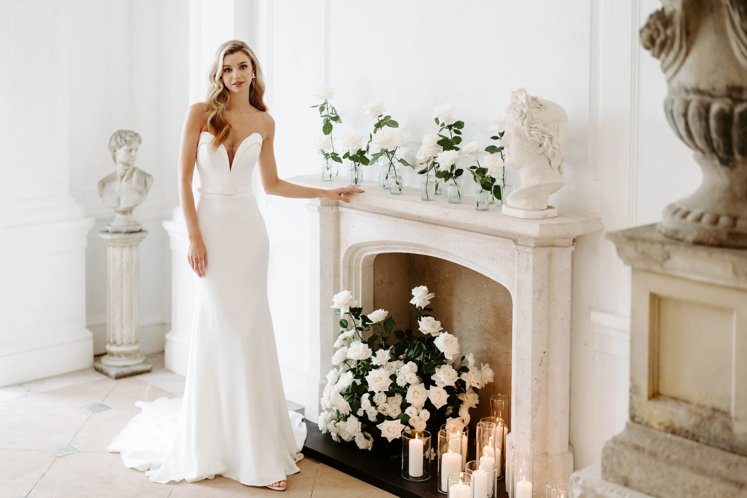 A woman in a white strapless wedding gown standing by a white fireplace decorated with white roses and candles, in a bright, elegant room.