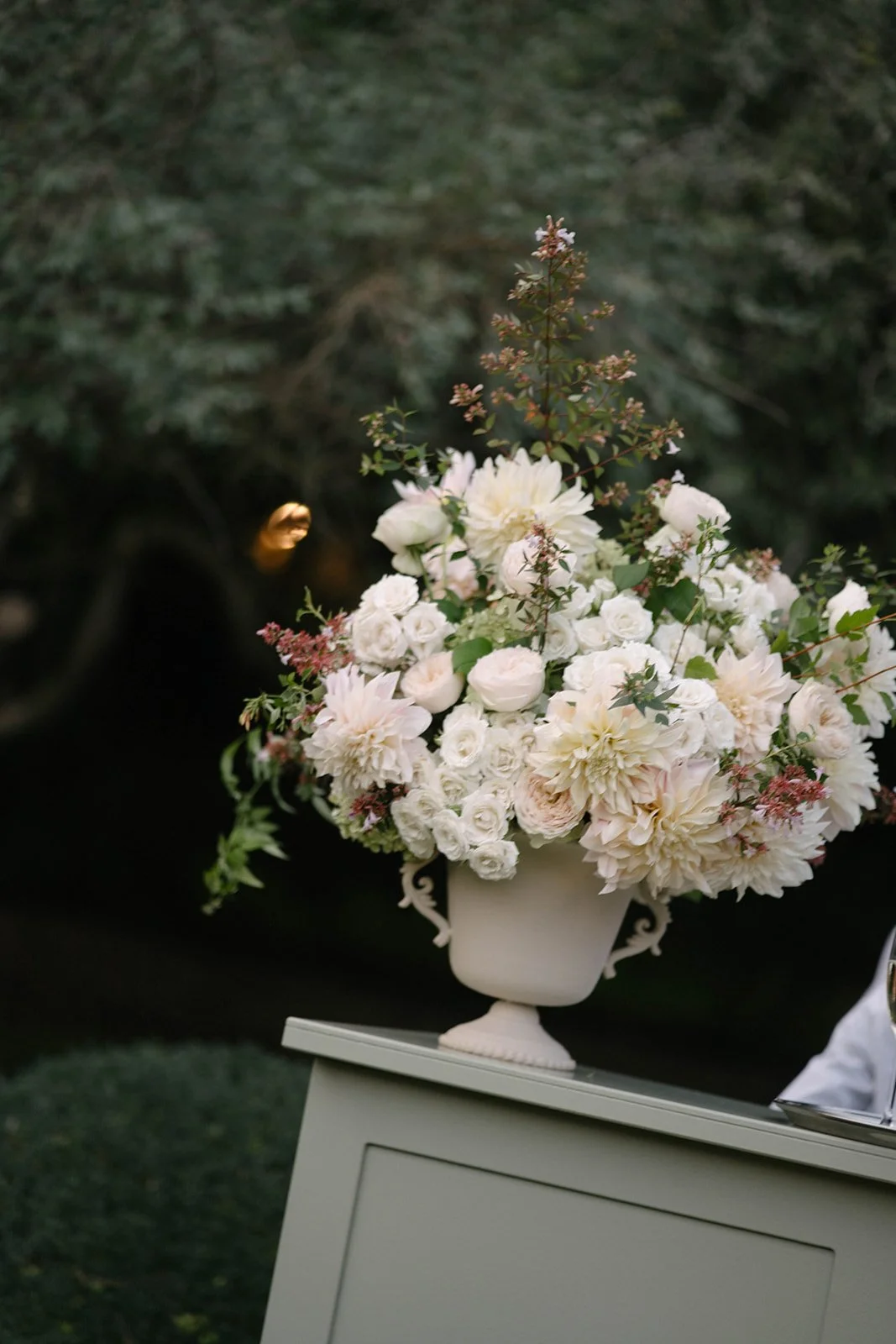 A white  Floral urn placed on a bar outdoors - – luxury garden-style wedding flowers by The Botany House, Cornwell Manor wedding florist in Oxfordshire, specialising in romantic seasonal marquee florals