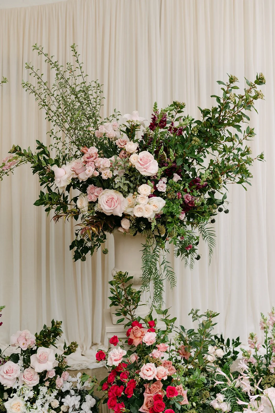 A large floral arrangement with pink and white roses, raspberry flowers, and green leaves in a white urn on a white pedestal, with a cream-colored curtain backdrop.  The Botany House Wedding Florist Kin House Planned by Get Wed