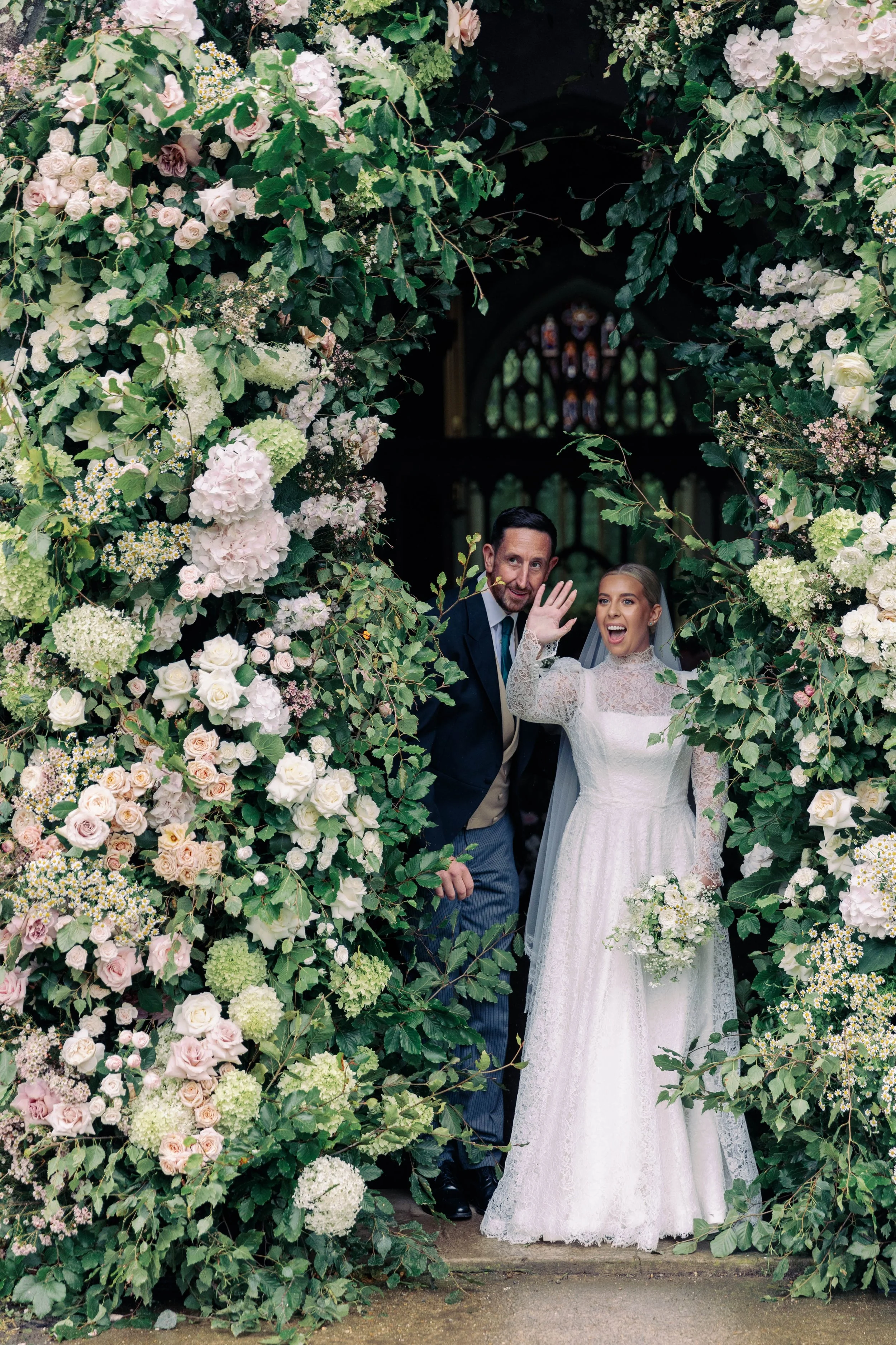 Bride in a white lace wedding dress and veil holding a bouquet of white flowers, standing next to a groom in a dark suit, white shirt, and blue tie, waving and smiling, all framed by a floral arch of inside a church with stained glass windows in the