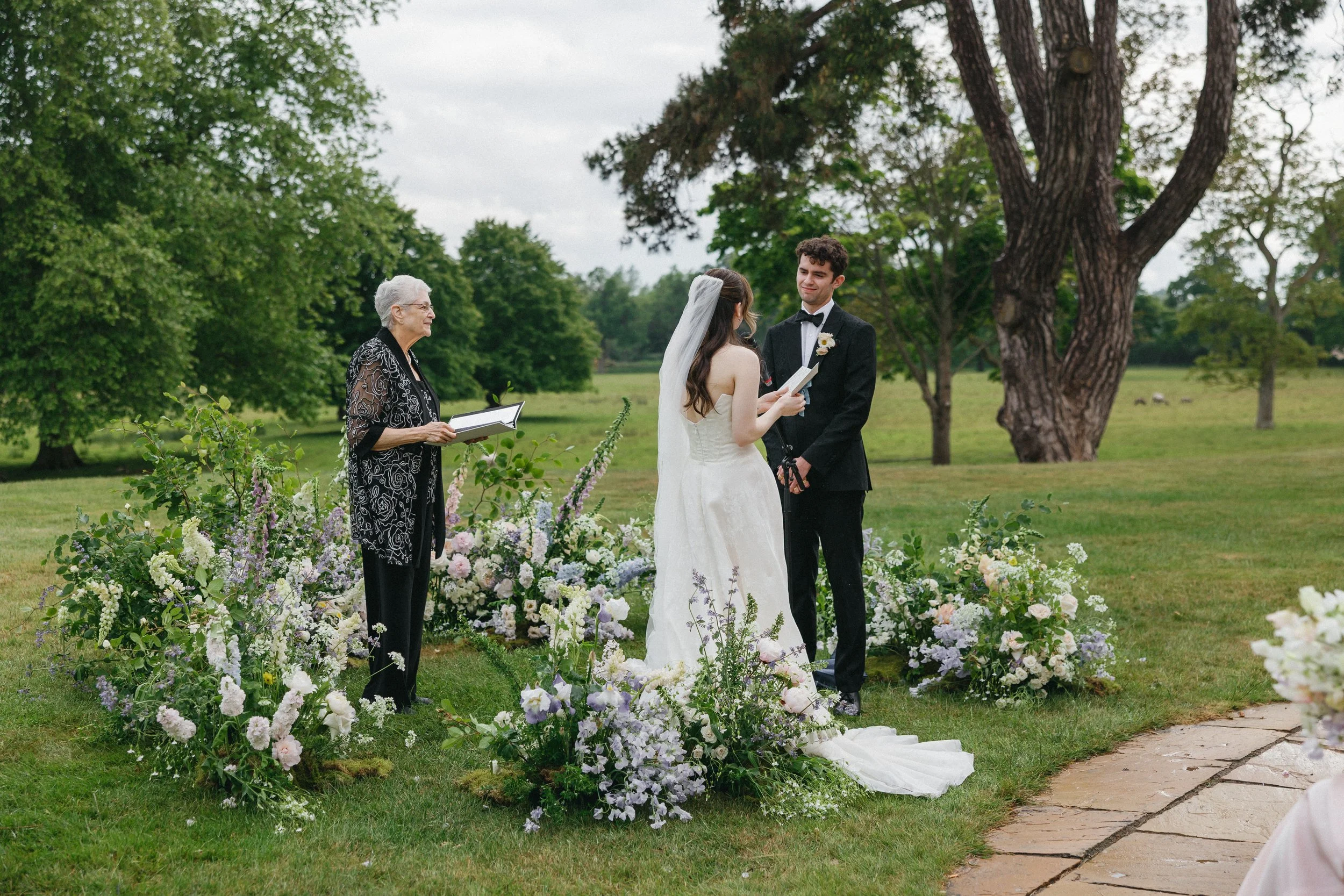 A bride and groom exchanging vows in an outdoor wedding ceremony with an officiant, surrounded by floral arrangements on a grassy area with trees in the background.