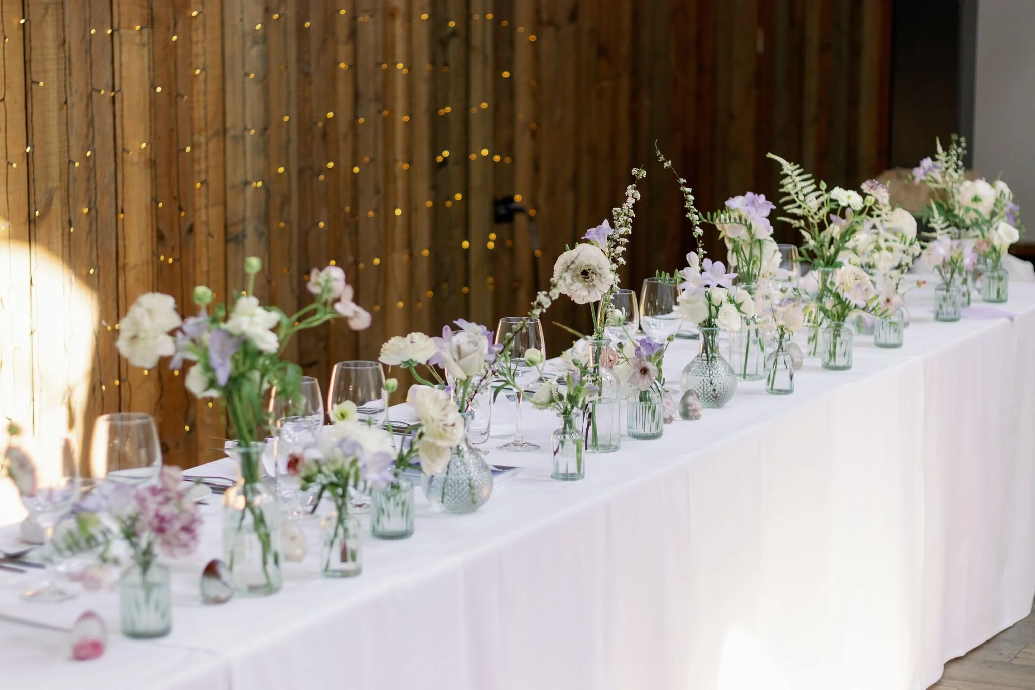 Elegant table decorated with white and soft pastel floral arrangements in glass vases, set against a wooden wall with fairy lights, for a wedding or special event.