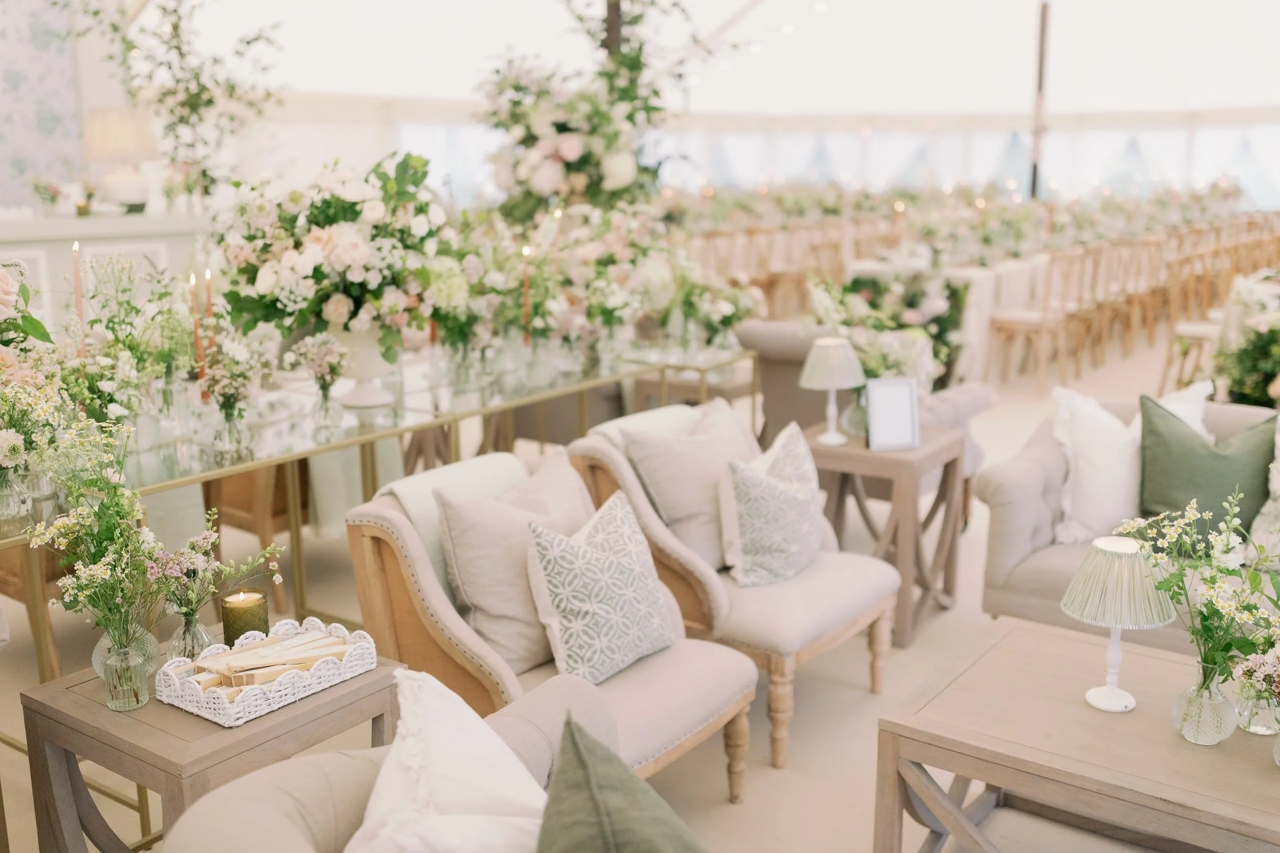 Elegant wedding seating area with neutral sofas, pastel throw pillows, small wooden side tables with vases of flowers, candles, and soft lighting. In the background, long tables decorated with abundant floral arrangements and candles.