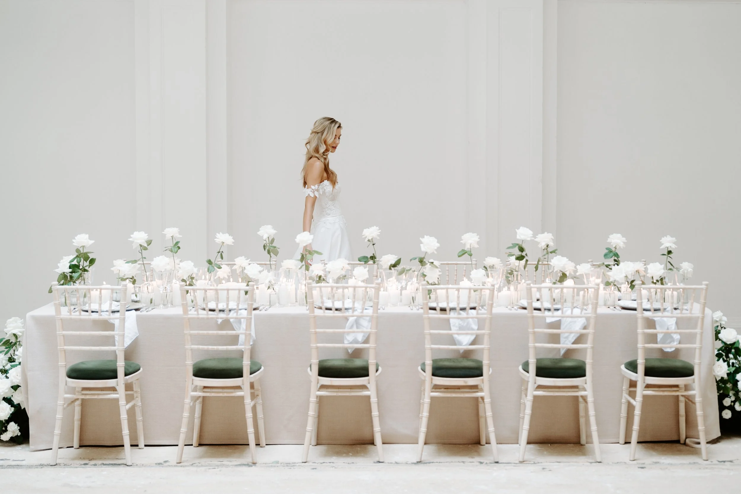 A woman in a white wedding dress standing behind a long, white table decorated with white flowers and candles, set up for a wedding reception.