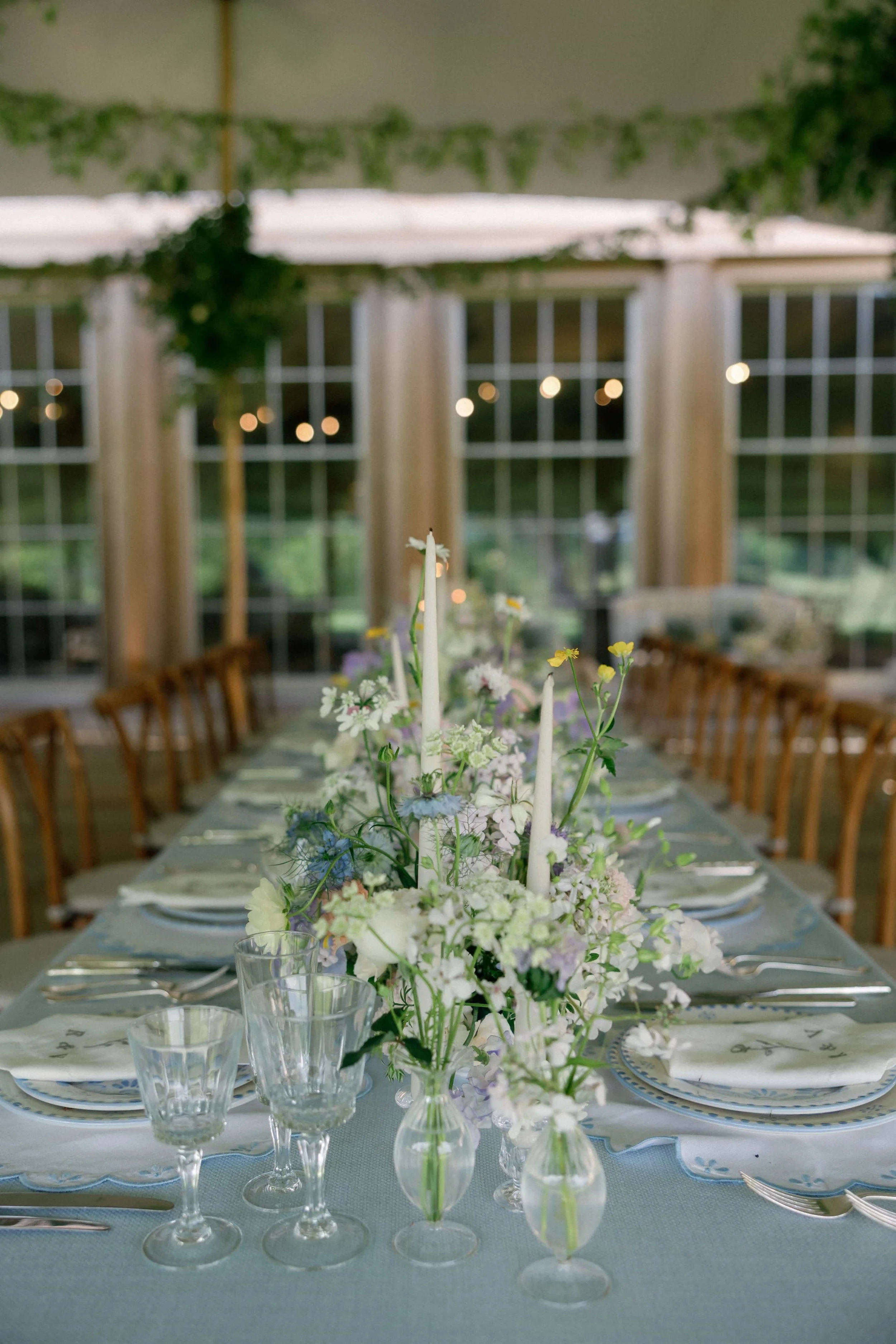 A decorated dining table with floral centerpiece, candles, glassware, and dinnerware set in a bright, airy room with large windows and string lights. Wilderness Reserve, Suffolk Florals By The Botany House