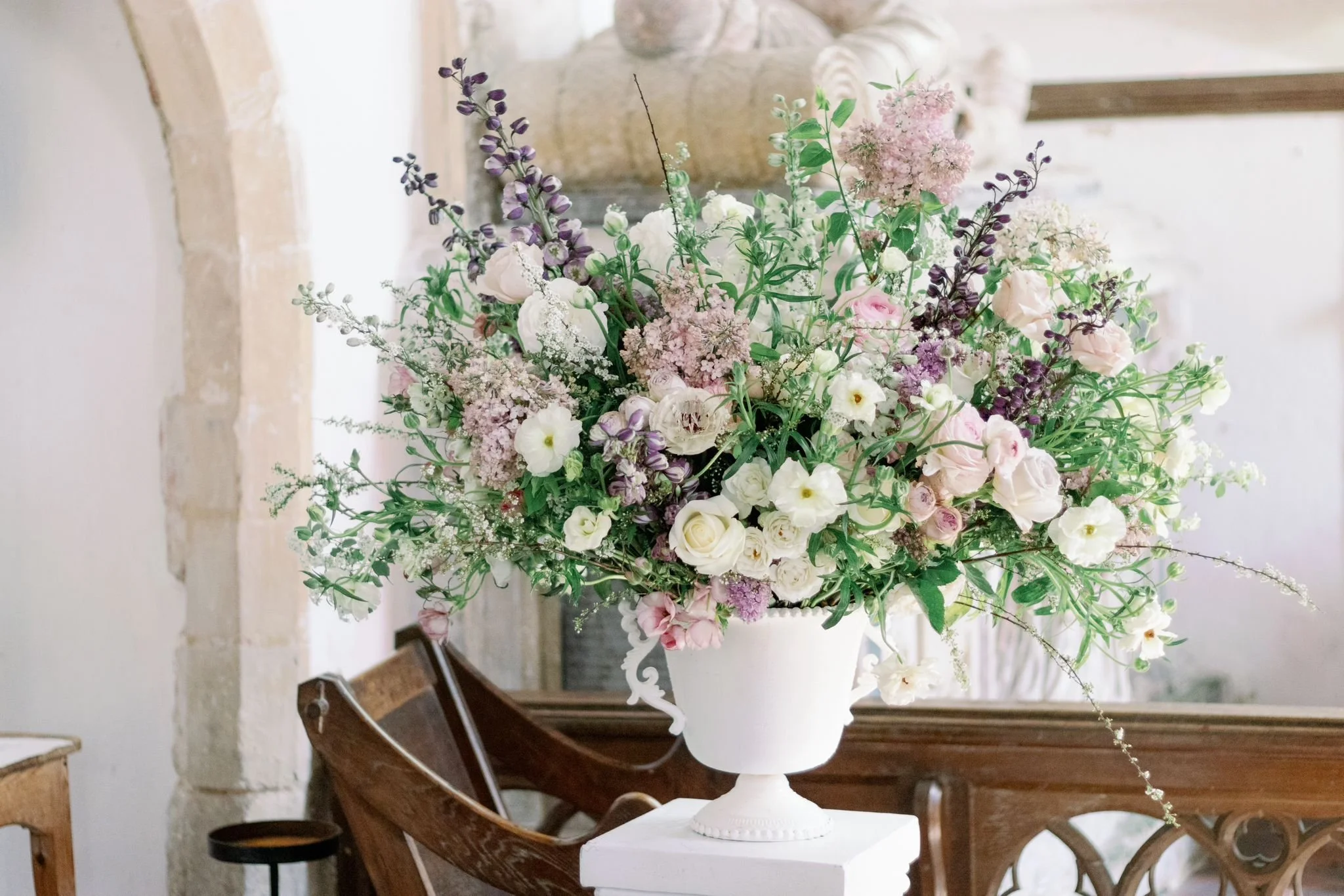 A large bouquet of white, pink, purple, and lavender flowers in a white vase on a white pedestal, placed on a wooden table with a rustic background.