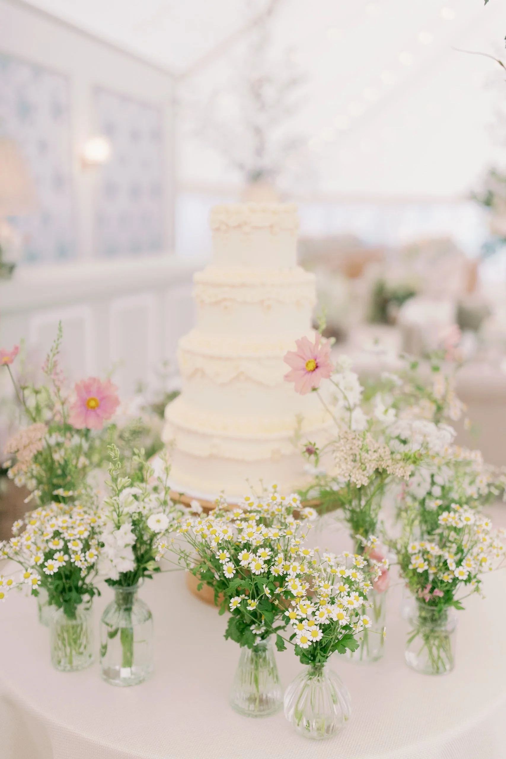 A white, multi-layered wedding cake on a table decorated with small glass vases filled with white and pink flowers.