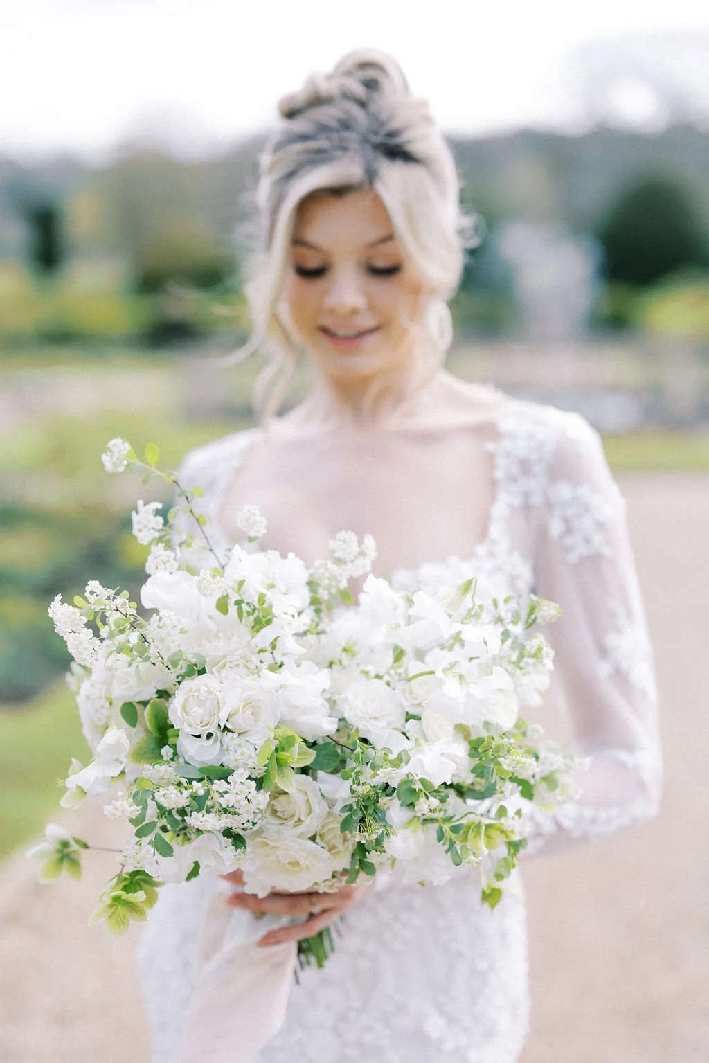 A bride holding a large bouquet of white flowers, outdoors on a bright day with greenery in the background.