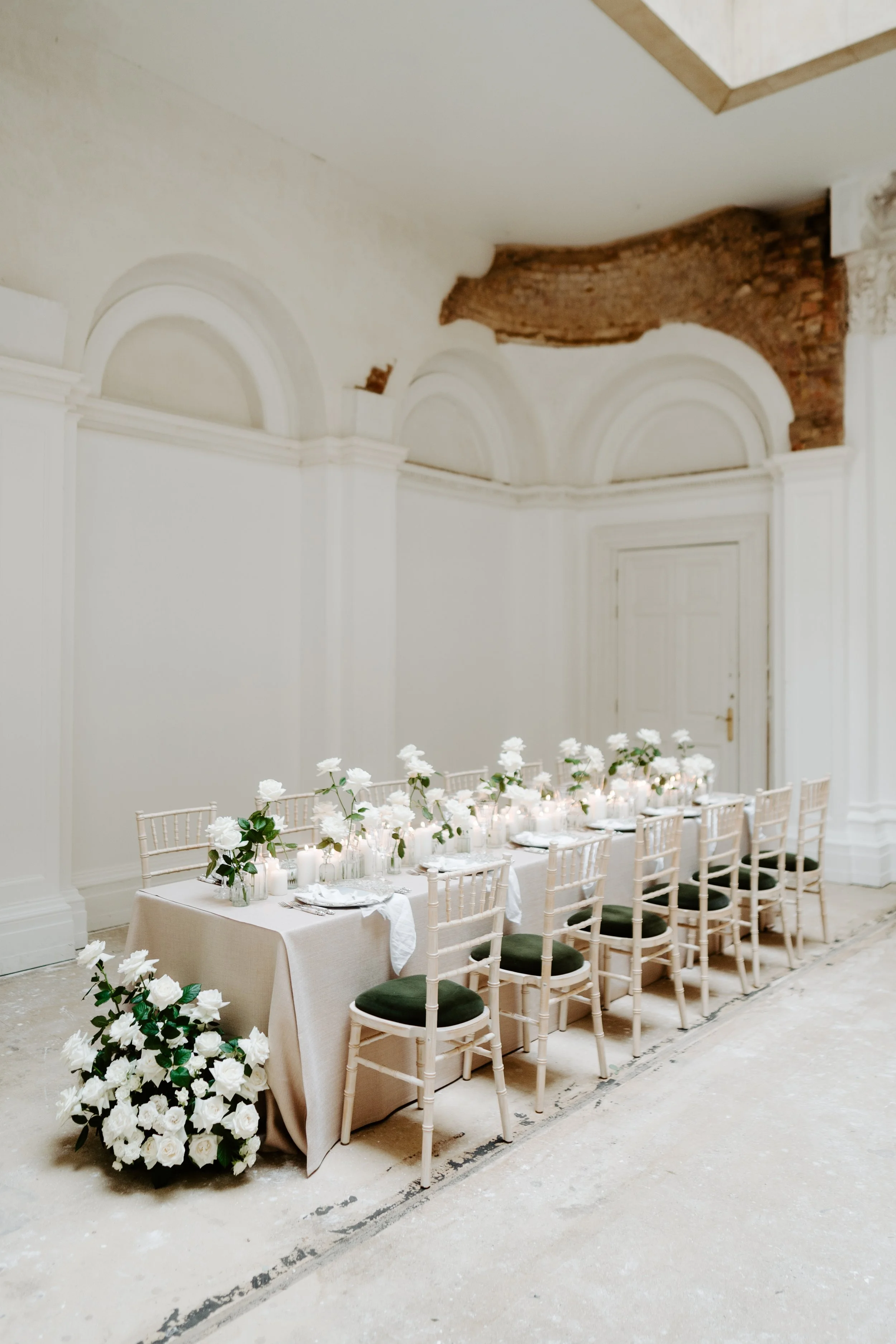 Long rectangular dining table decorated with white roses and candles, surrounded by white chairs with black cushions, in a room with white walls and ornate arch designs.