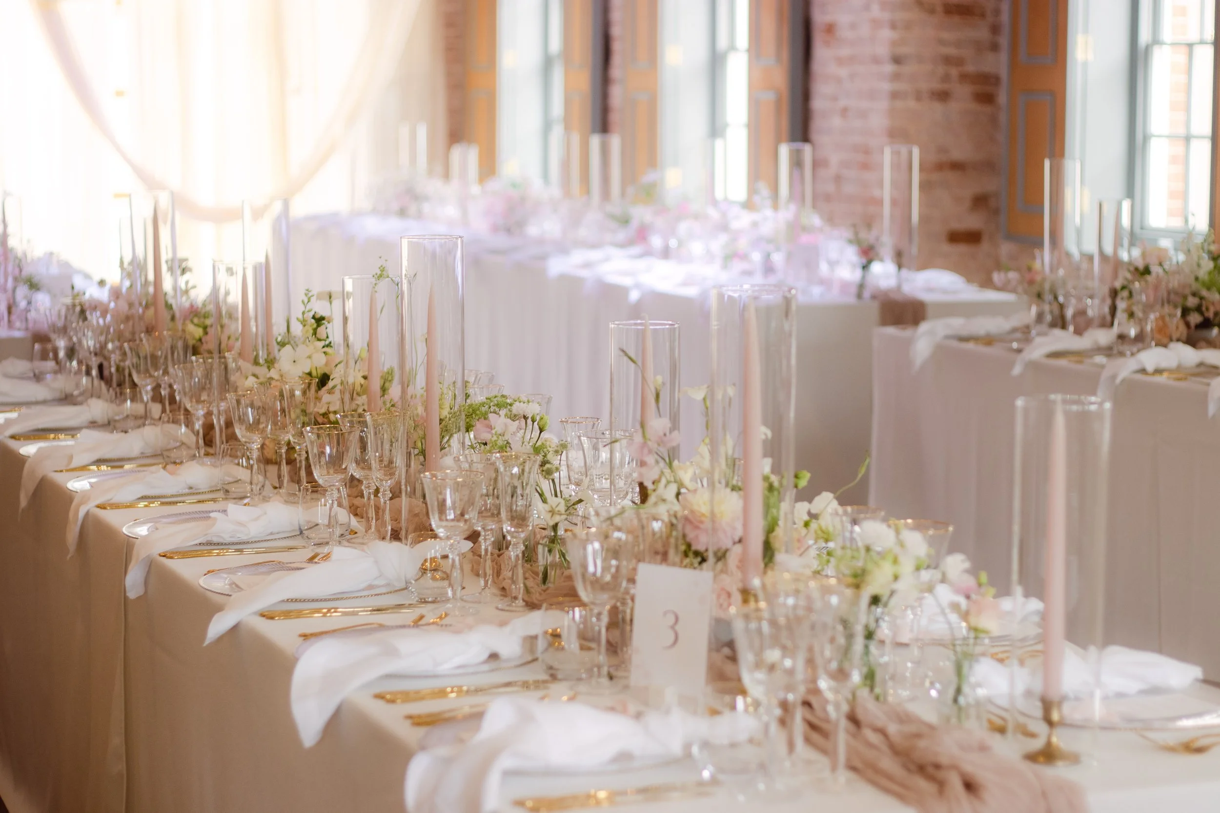Decorated banquet table at a wedding reception with pink candles, white flowers, and gold cutlery in a bright room with large windows.