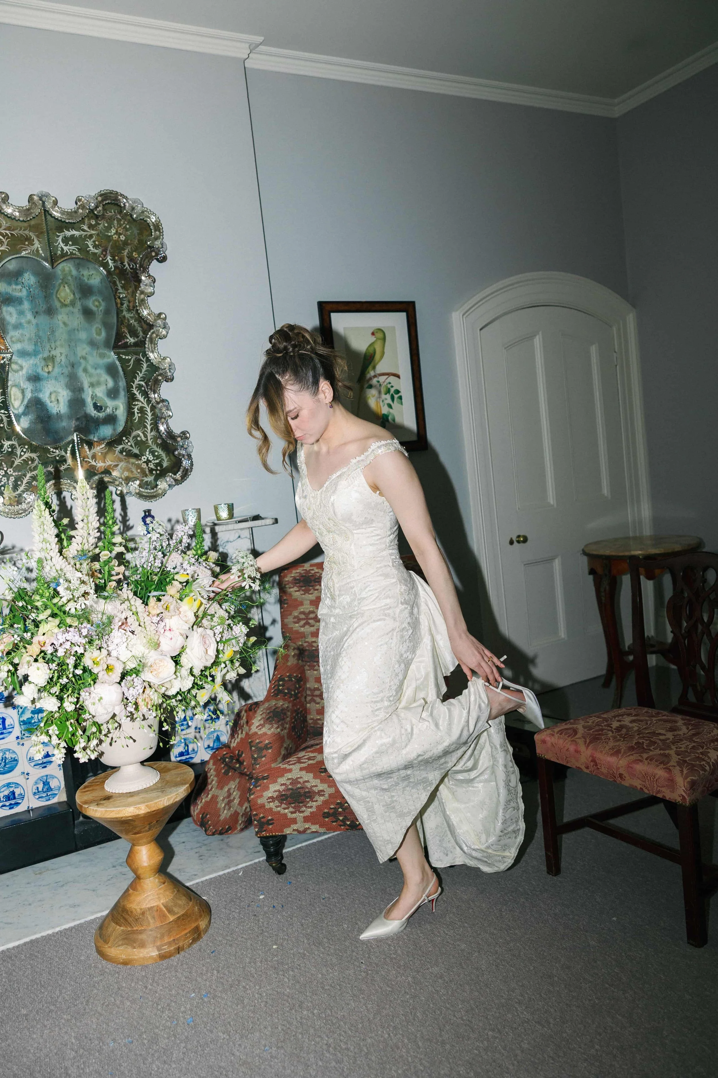A woman in a wedding dress fixing her shoe in a room decorated with flowers and artwork.