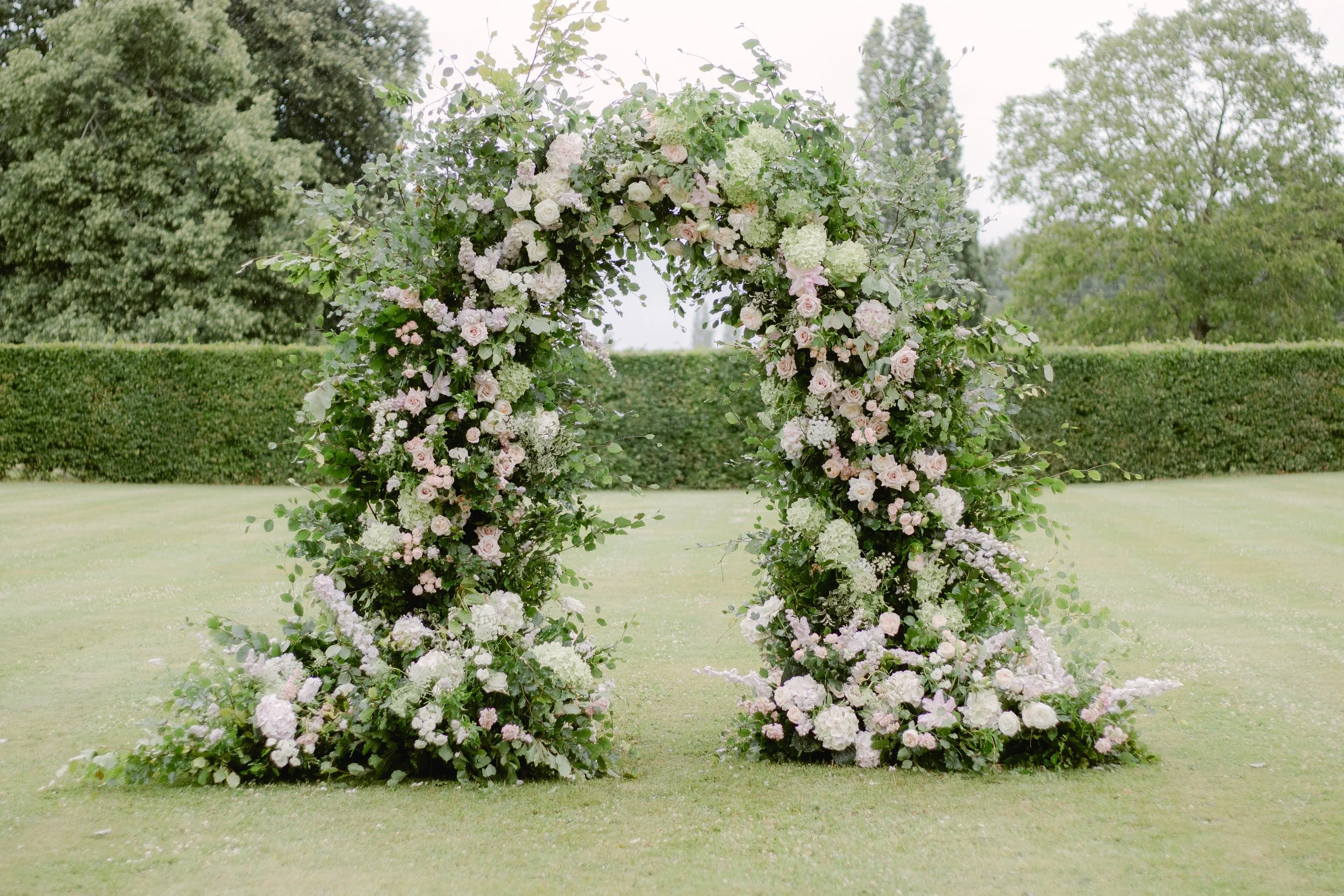 Large floral wedding arch decorated with white and light pink roses, hydrangeas, and green foliage on a grassy outdoor lawn with trees and hedges in the background.