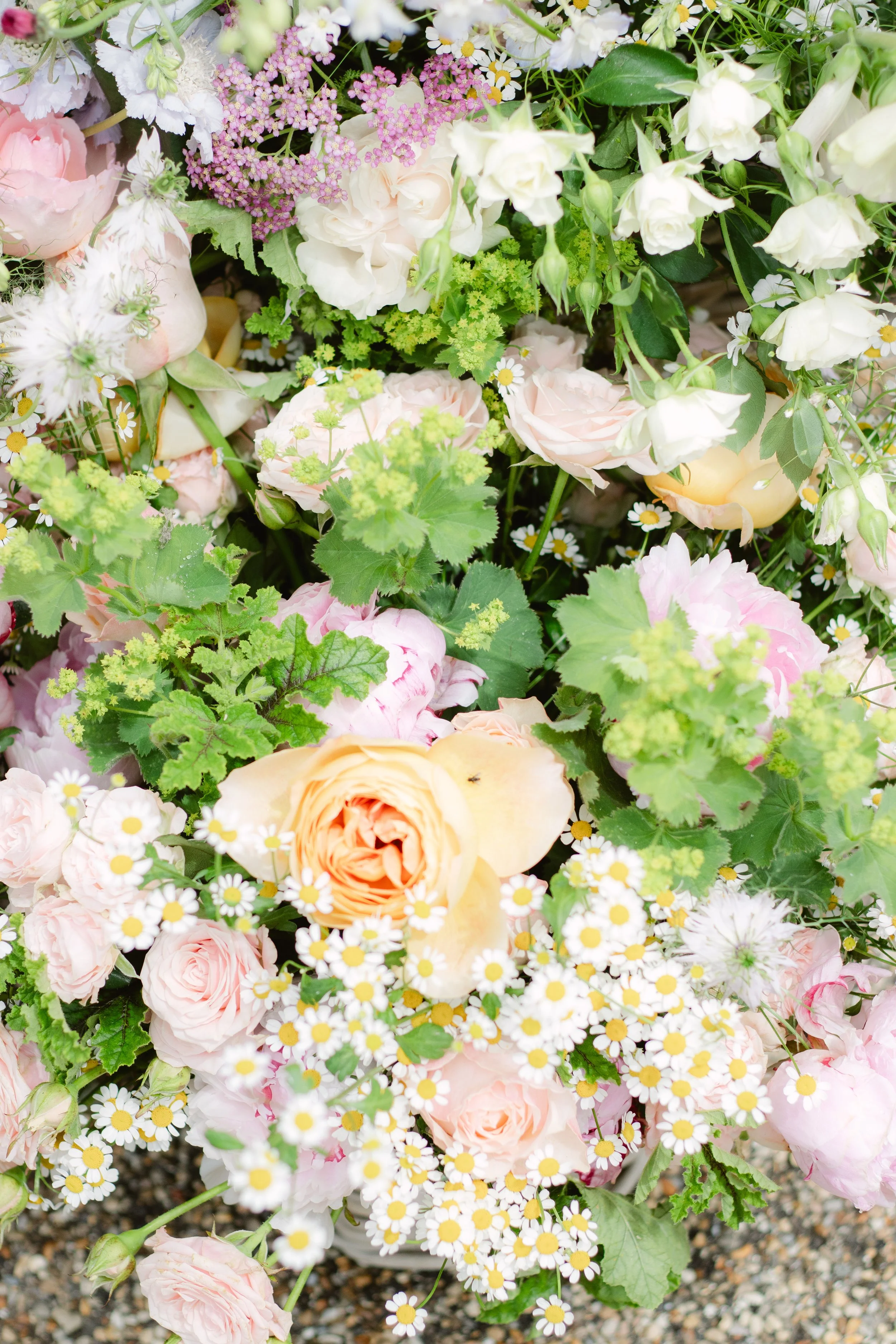 A lush arrangement of various flowers, including pale pink, peach, and white roses, white bell-shaped flowers, small white daisies with yellow centers, and green leafy foliage.