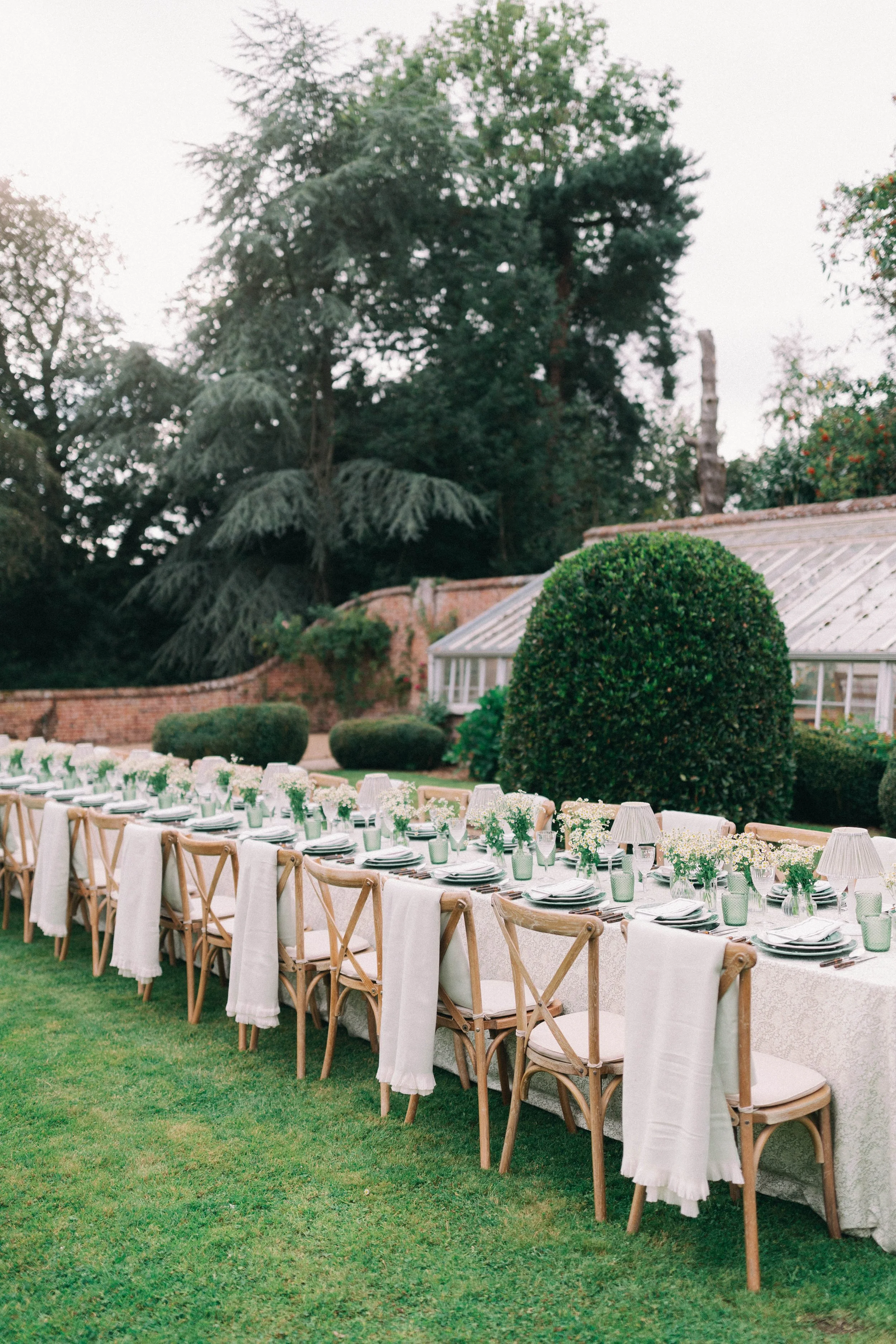 Long outdoor dining table set with floral tablecloth, plates, napkins, glasses, and small flower arrangements, surrounded by wooden chairs with white blankets draped over the backs, in a garden with trees and shrubs. Hosted near Holt in Norfolk