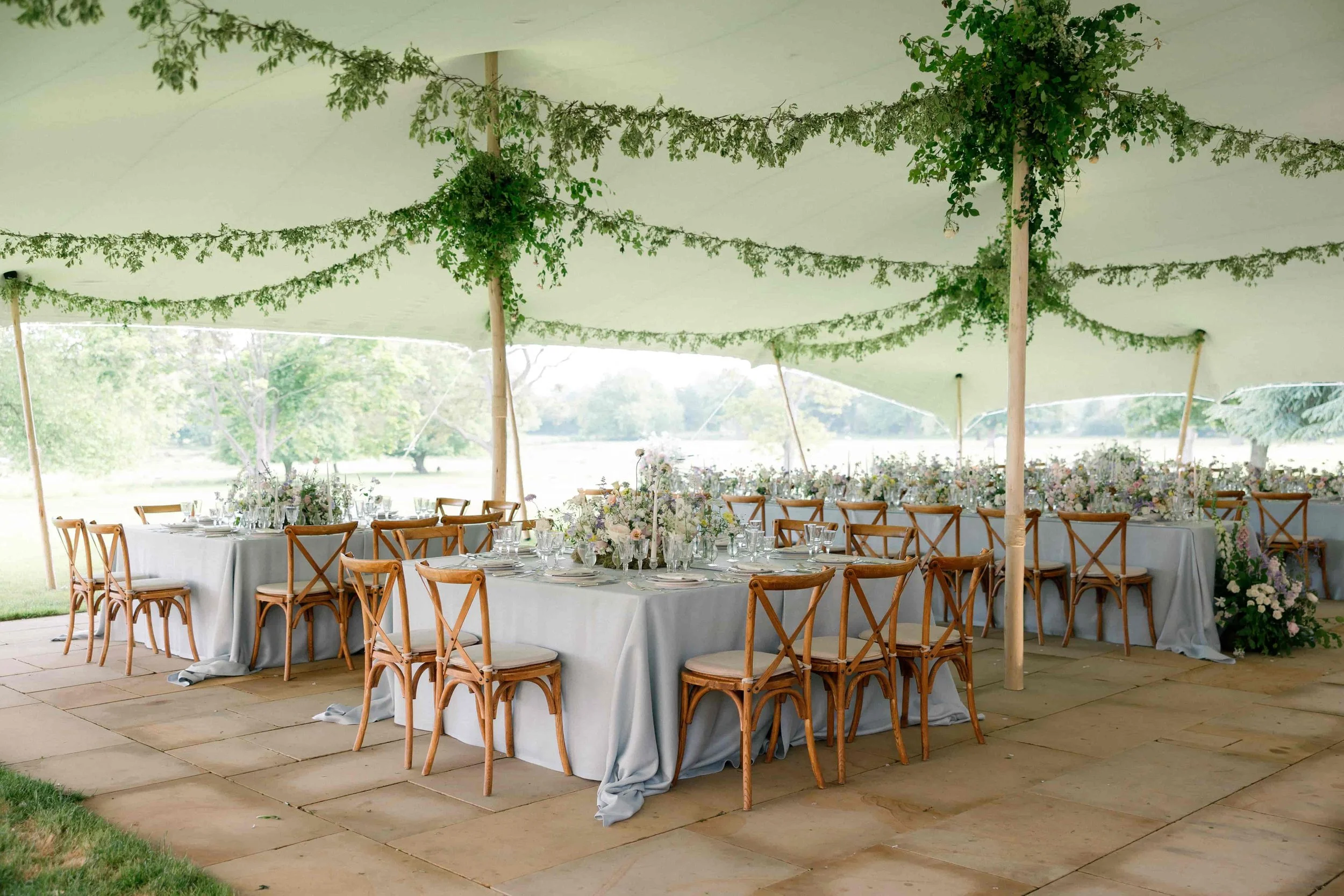 Wedding reception setup with long tables covered in white tablecloths, decorated with floral centerpieces, under a large white tent with green foliage hanging from the ceiling, overlooking a grassy outdoor area. The Botany House Florist at Wilderness