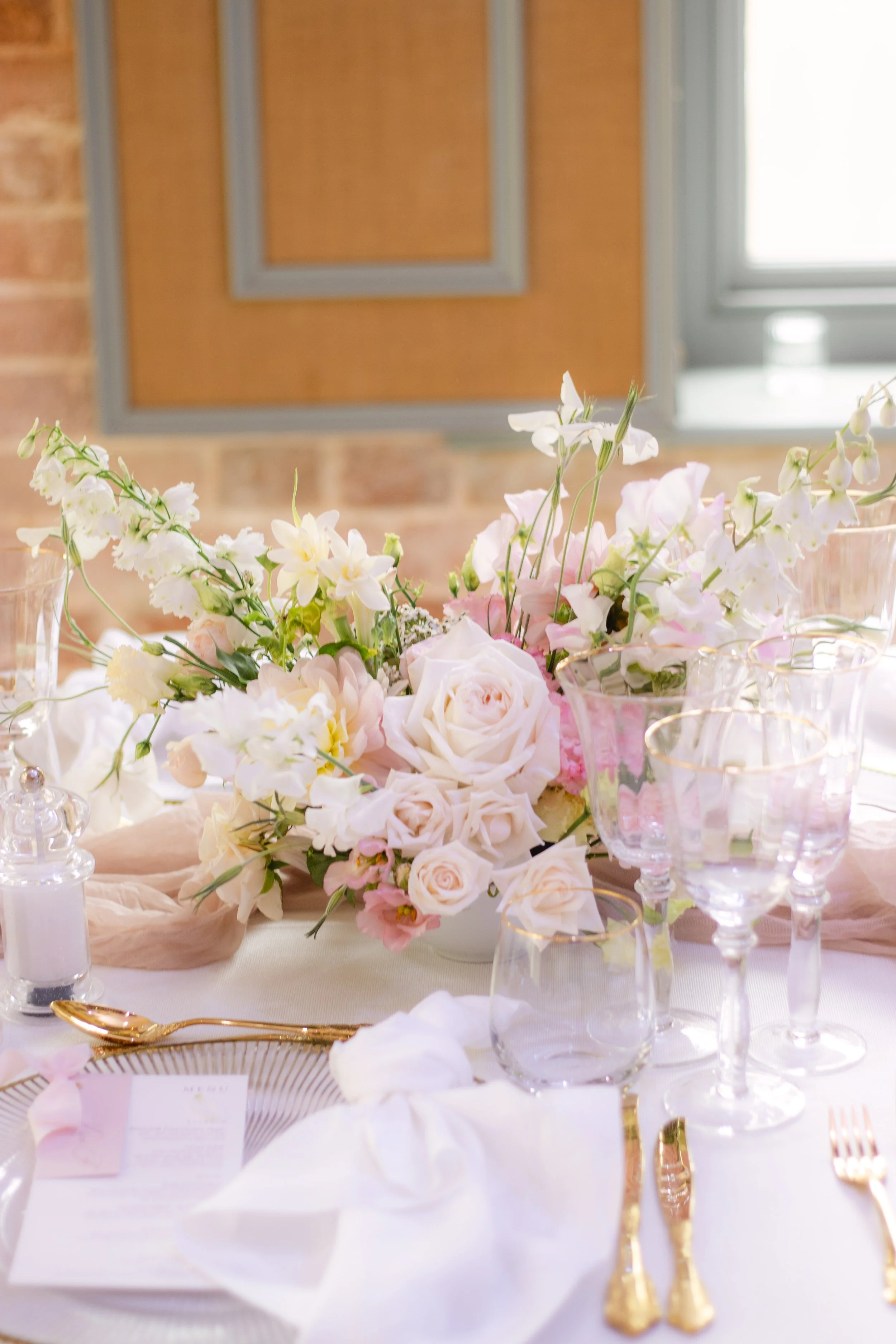 Elegant table setting with pink and white floral centerpiece, gold utensils, and glassware on a white tablecloth, near a window with brick wall background.