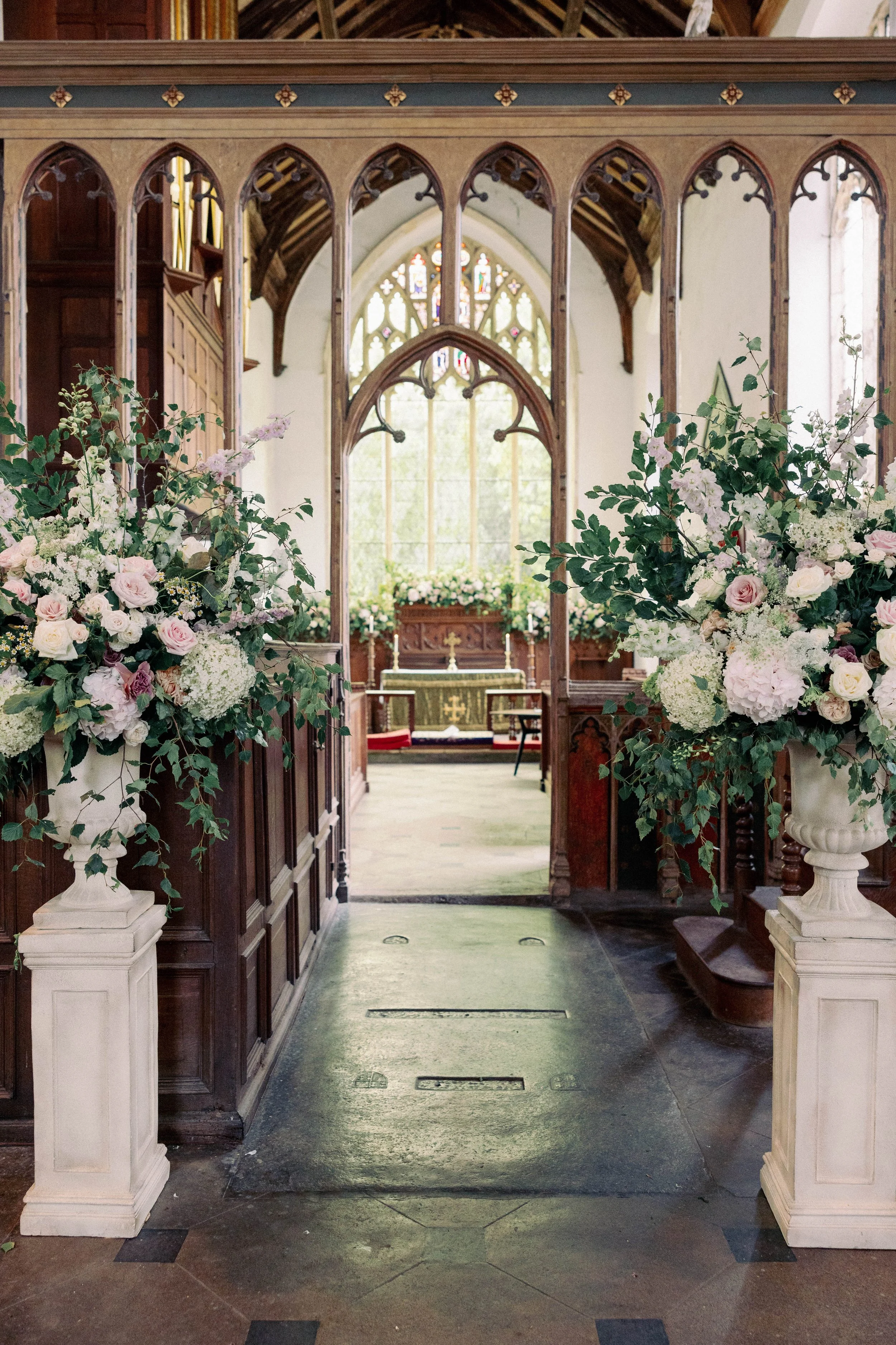 View of a church interior with wooden pews, floral arrangements in white urns on pedestals, and stained glass windows at the altar.