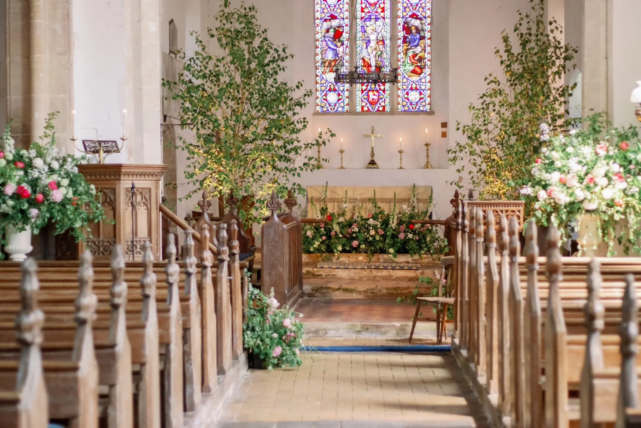 Interior of a church altar decorated with pink and white flowers, two small trees, and stained glass windows above the altar.