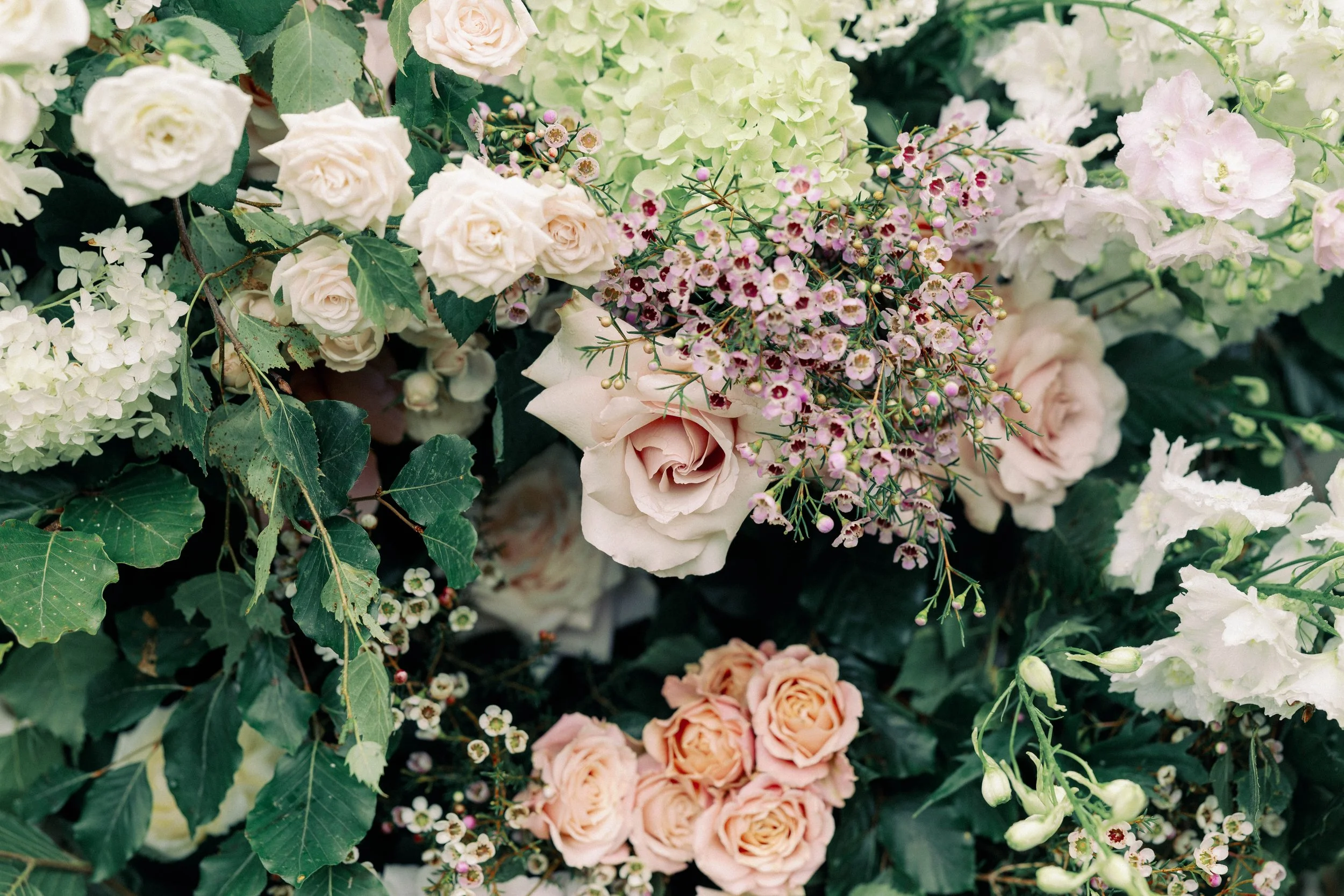 Various light-colored flowers, including white and pale pink roses, light green hydrangeas, and small pink and white blossoms, with green leaves surrounding them. Garden style florals in Norfolk