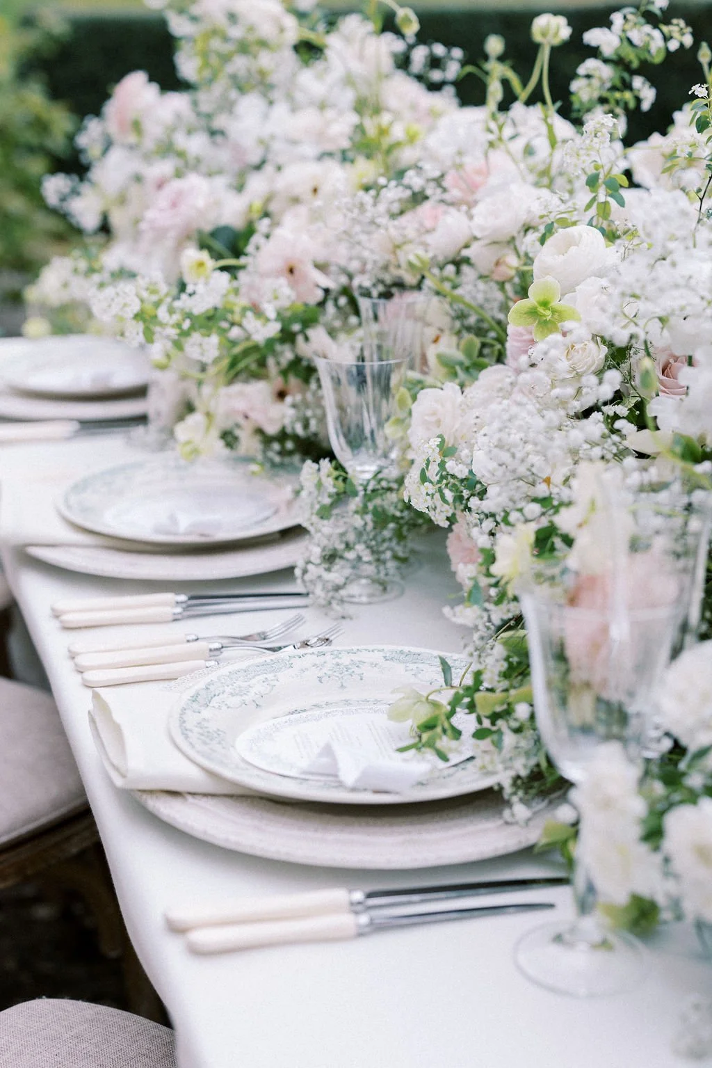 Elegant outdoor table setting with white plates, silverware, and tall glasses, decorated with a lush floral centerpiece of white and pastel pink flowers.