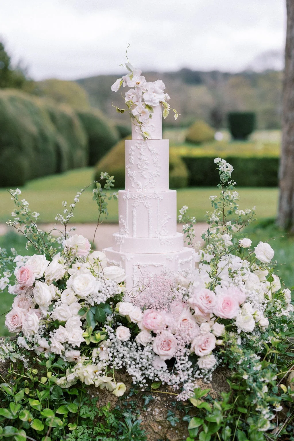 A tall, white wedding cake with four tiers decorated with floral icing details, topped with white flowers and greenery, surrounded by an arrangement of white and pink roses, baby's breath, and greenery, outdoors on a grassy area with blurred green hi