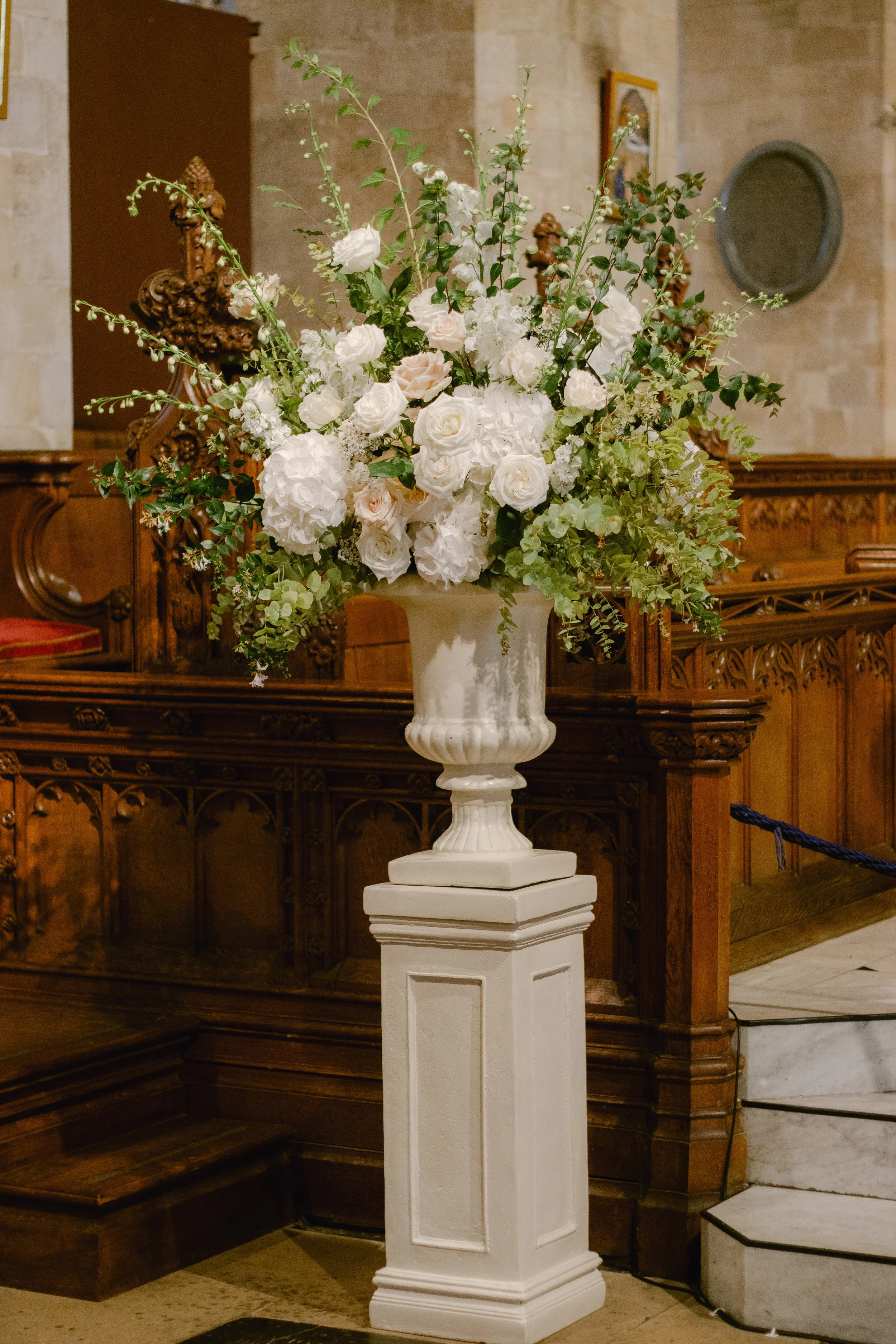 A large floral arrangement of white roses and hydrangeas on a white pedestal in a church with wooden pews and stone walls.