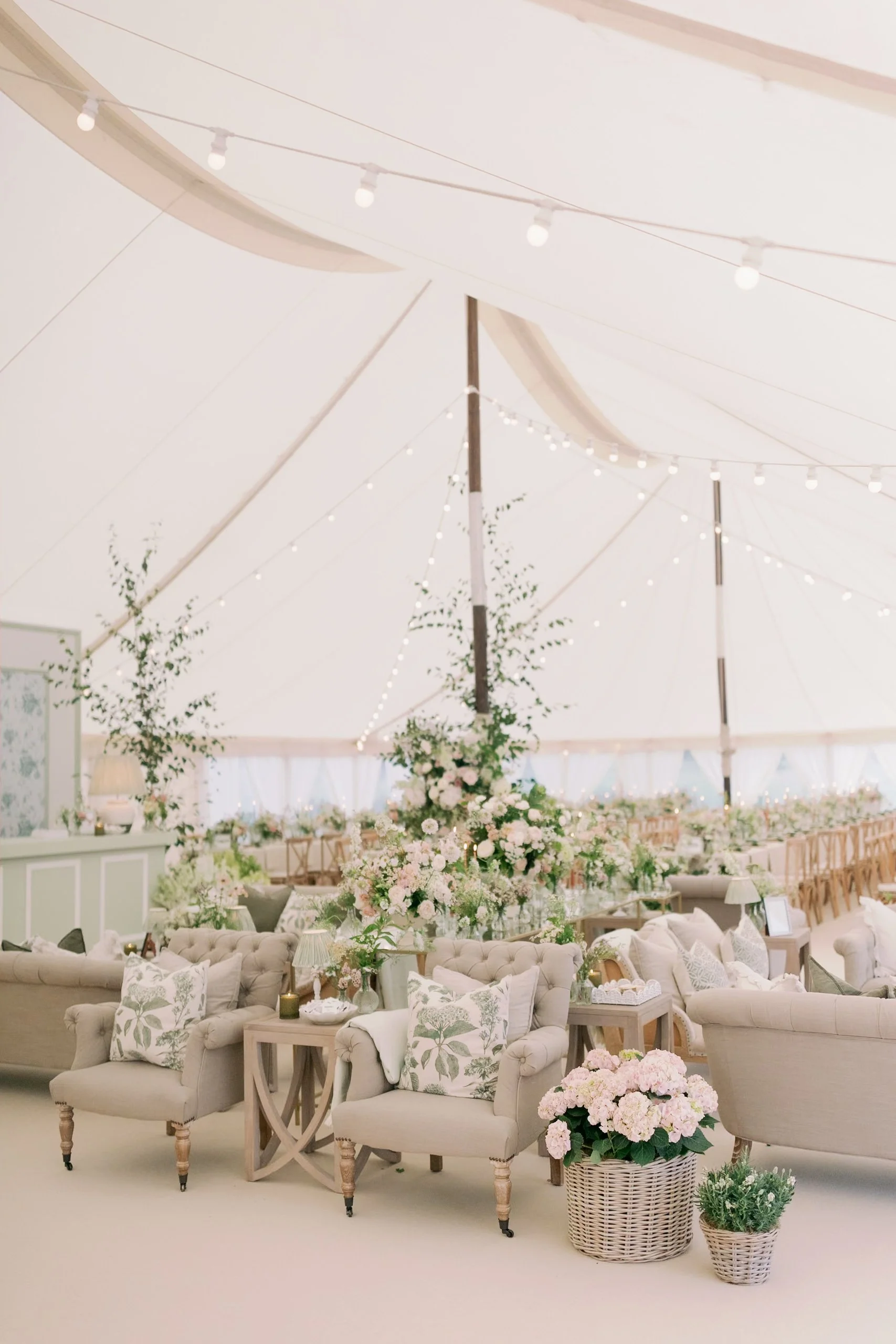 Elegant wedding reception in a large white marquee with beige sofas, floral arrangements, string lights, and wooden chairs.