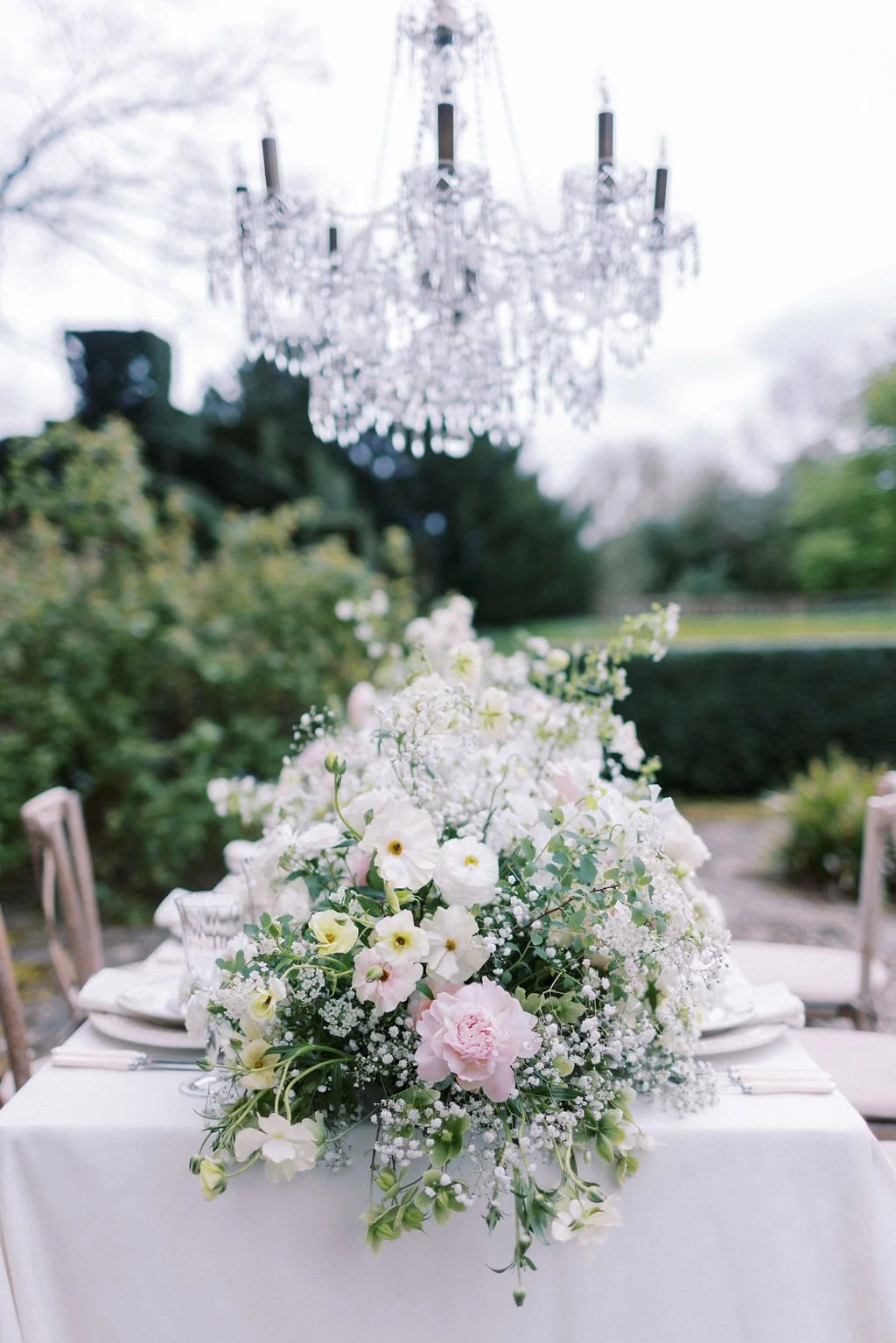 Elegant outdoor table setup with a large bouquet of white and pale pink flowers on a white tablecloth, with a crystal chandelier hanging above, surrounded by lush greenery and trees.