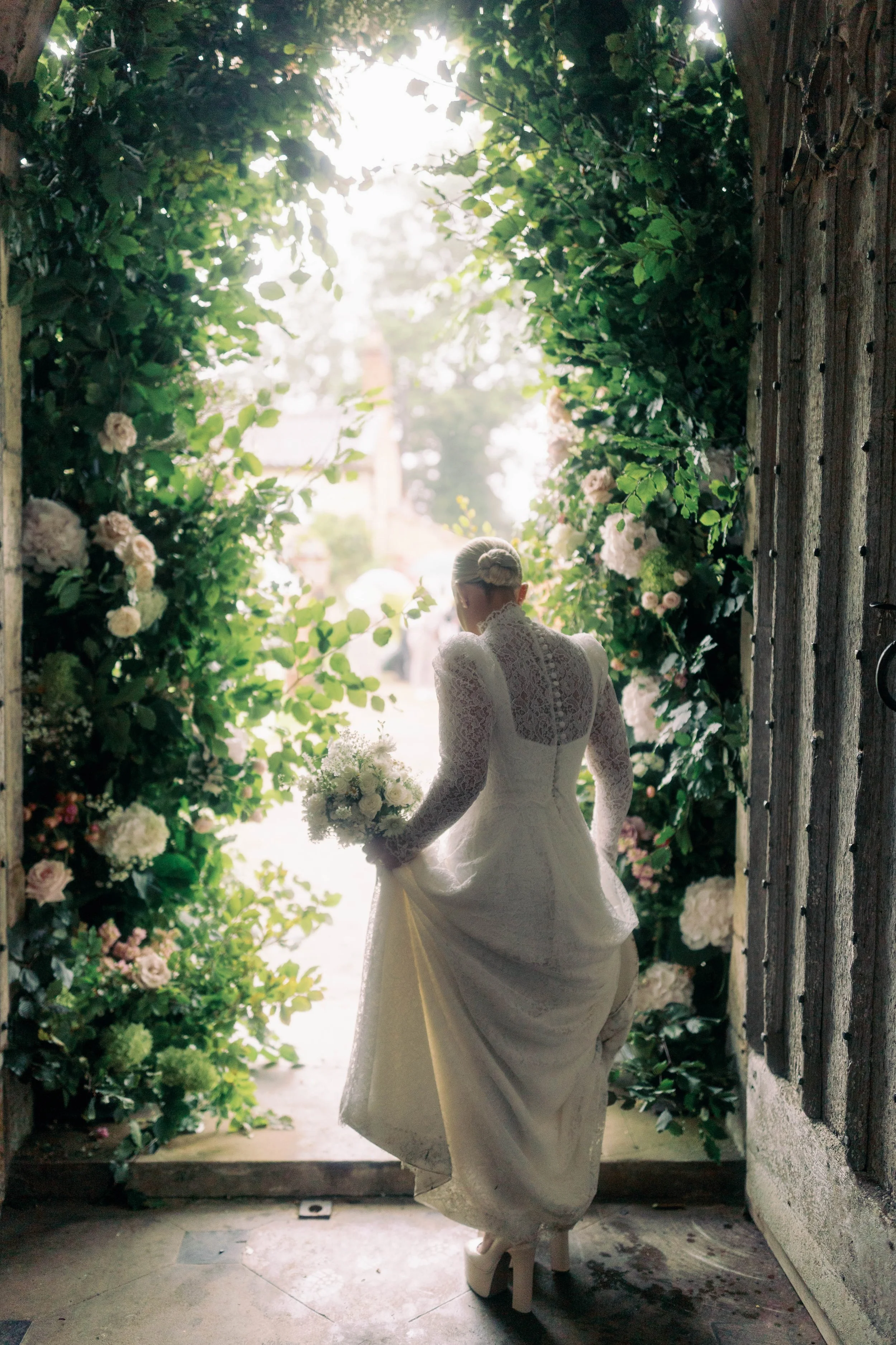 A bride in a lace wedding dress holding a bouquet, standing at the entrance of a garden archway decorated with flowers and greenery.